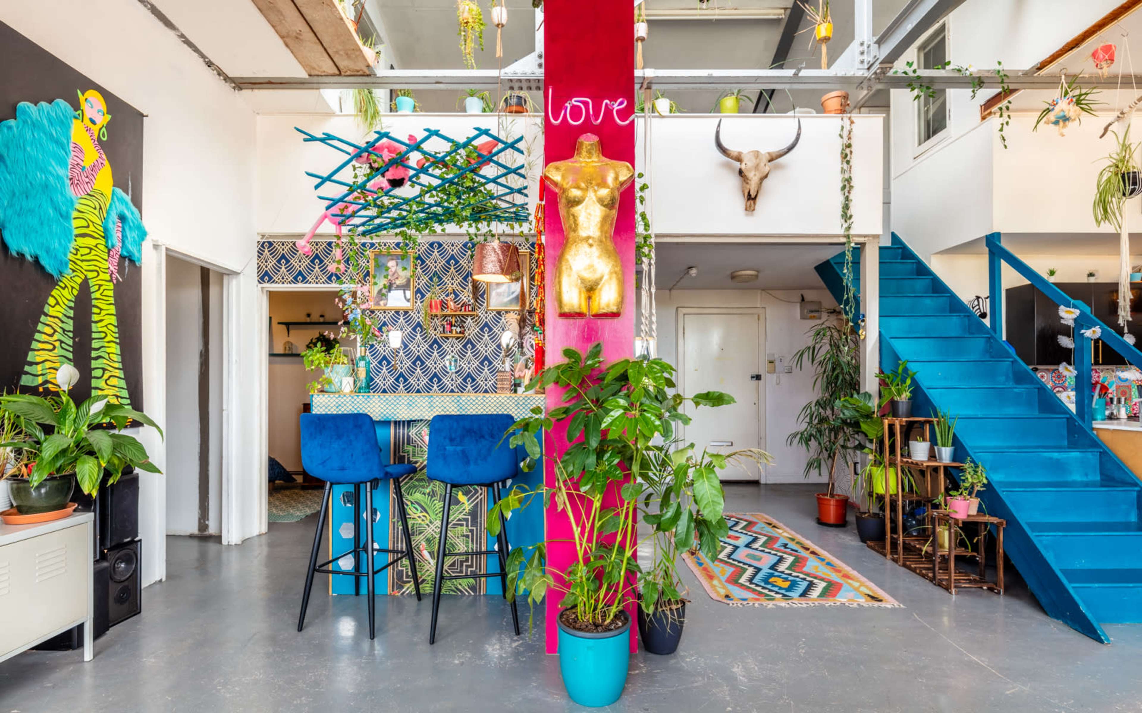 A colorful open-concept living space with a prominent pink and gold decorative column, blue barstools, and various plants surrounding a staircase.
