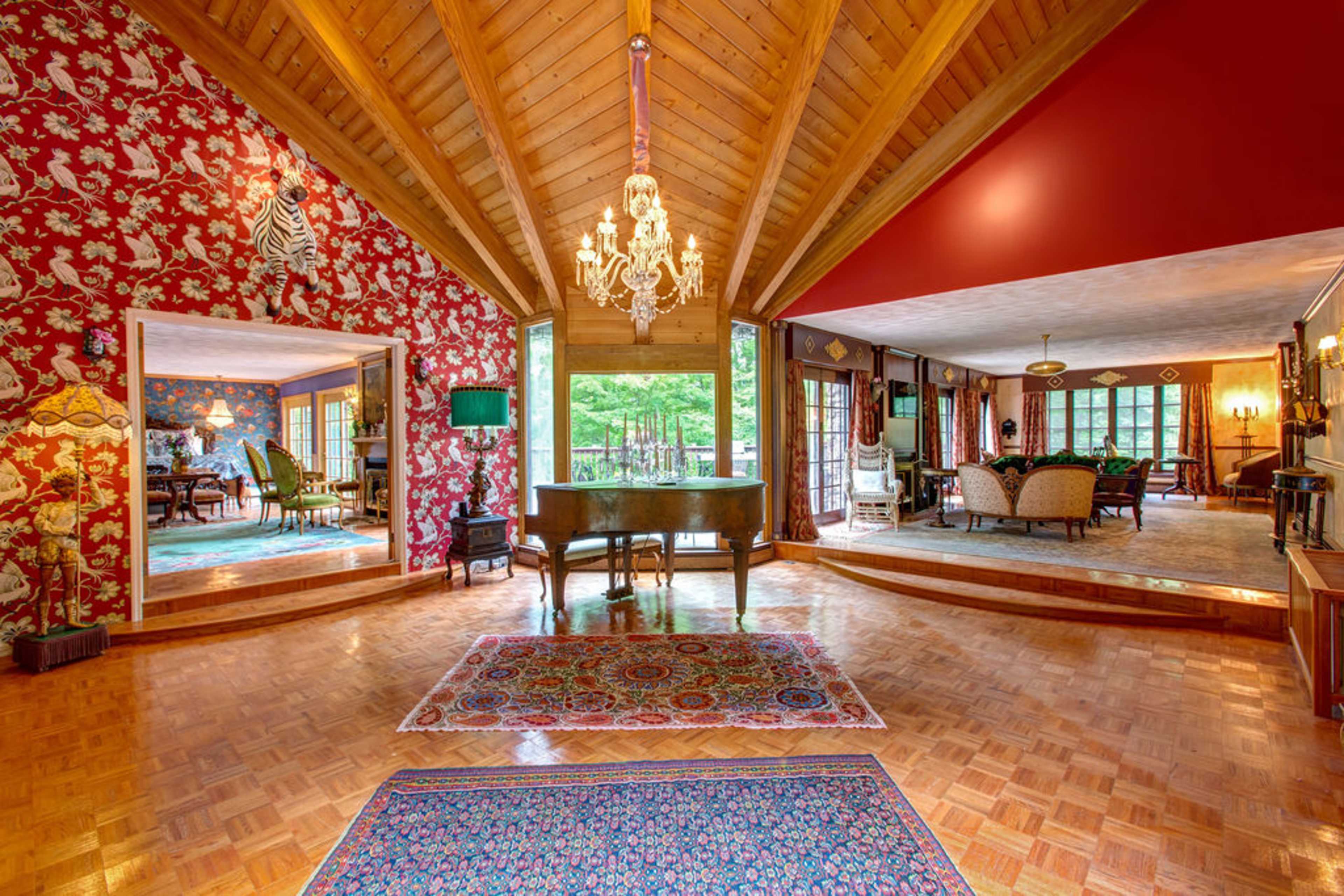 A spacious living area with high wooden ceilings, patterned wallpaper, a piano, and various seating arrangements, all illuminated by natural light.