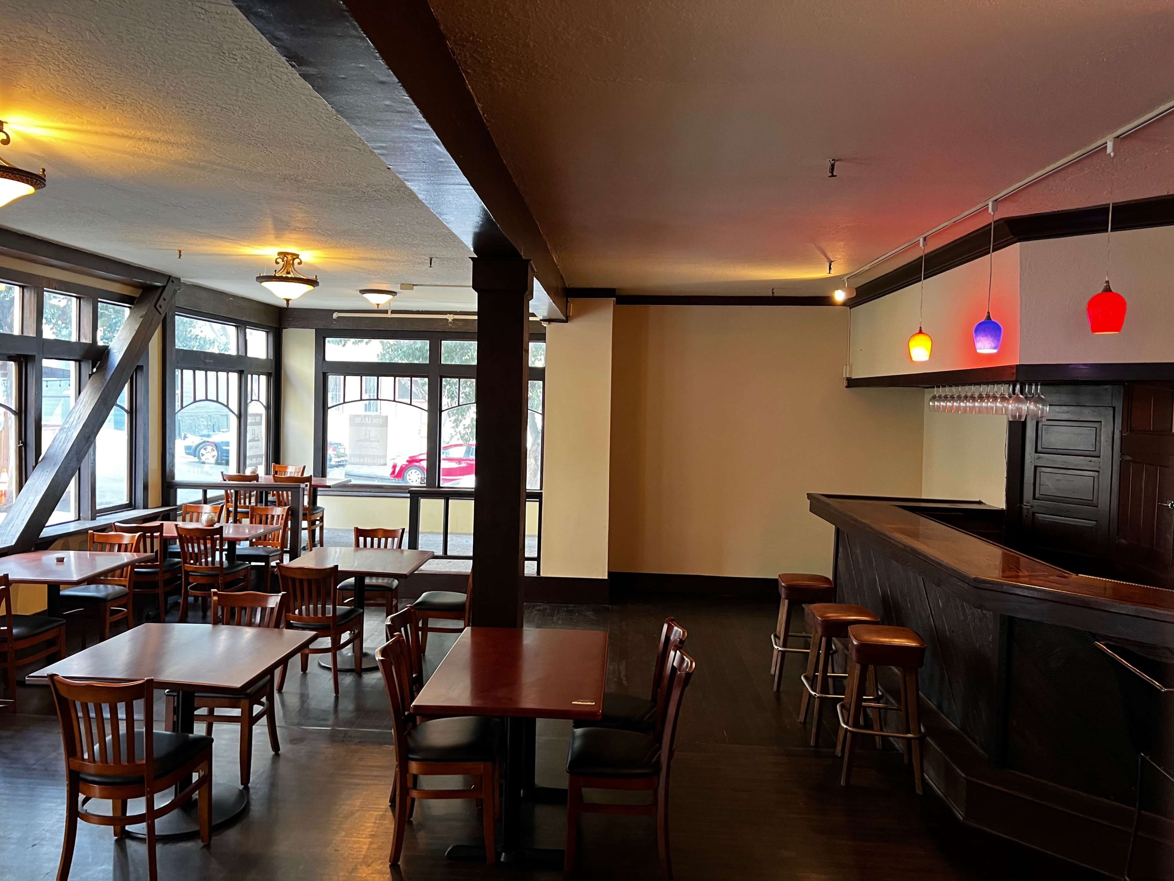 A spacious, empty restaurant interior featuring wooden tables and chairs, a bar with colorful pendant lights, and large windows allowing natural light.