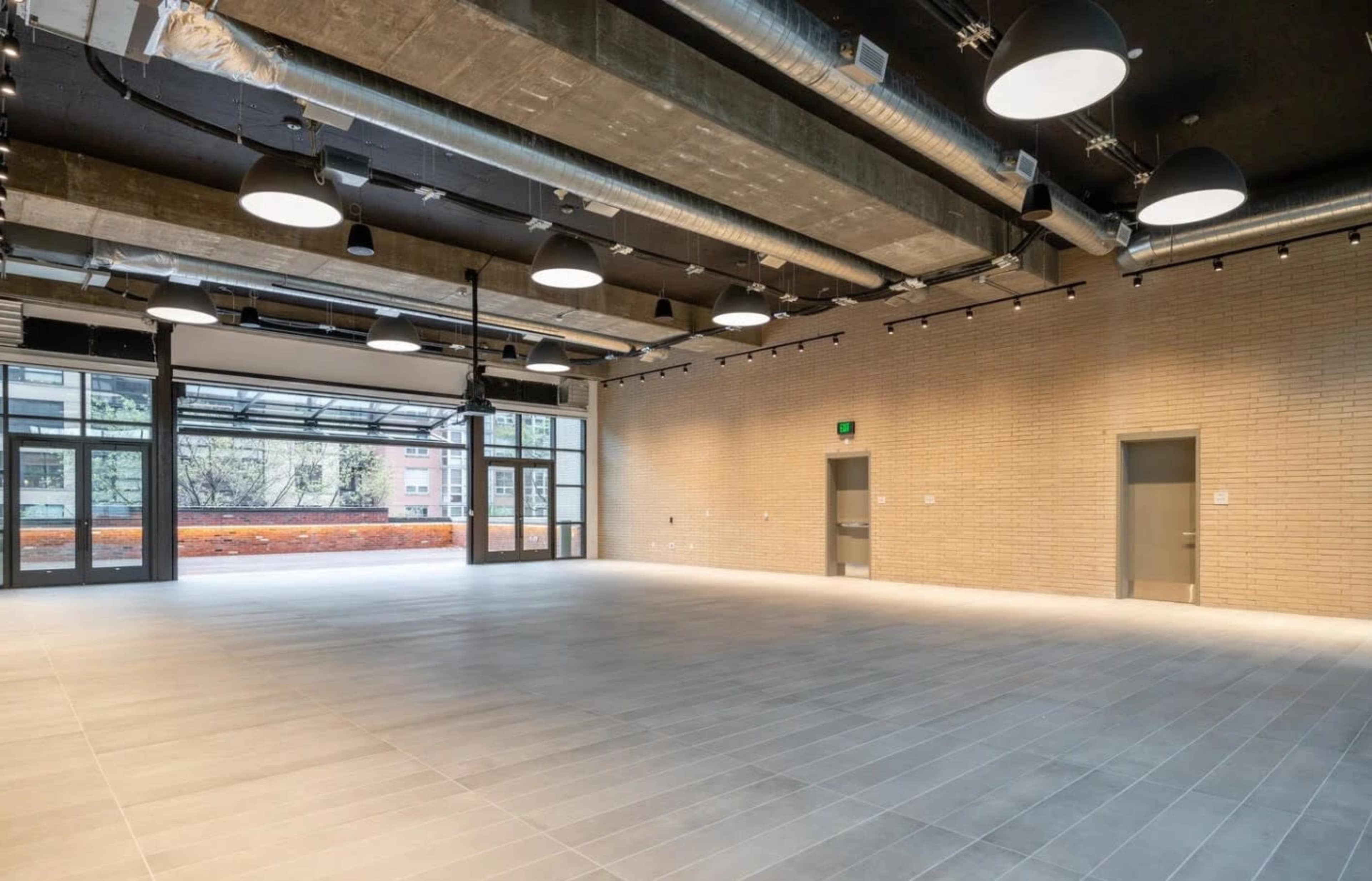 The image shows an empty, modern indoor space with large windows and high ceilings, featuring concrete walls and ample overhead lighting.