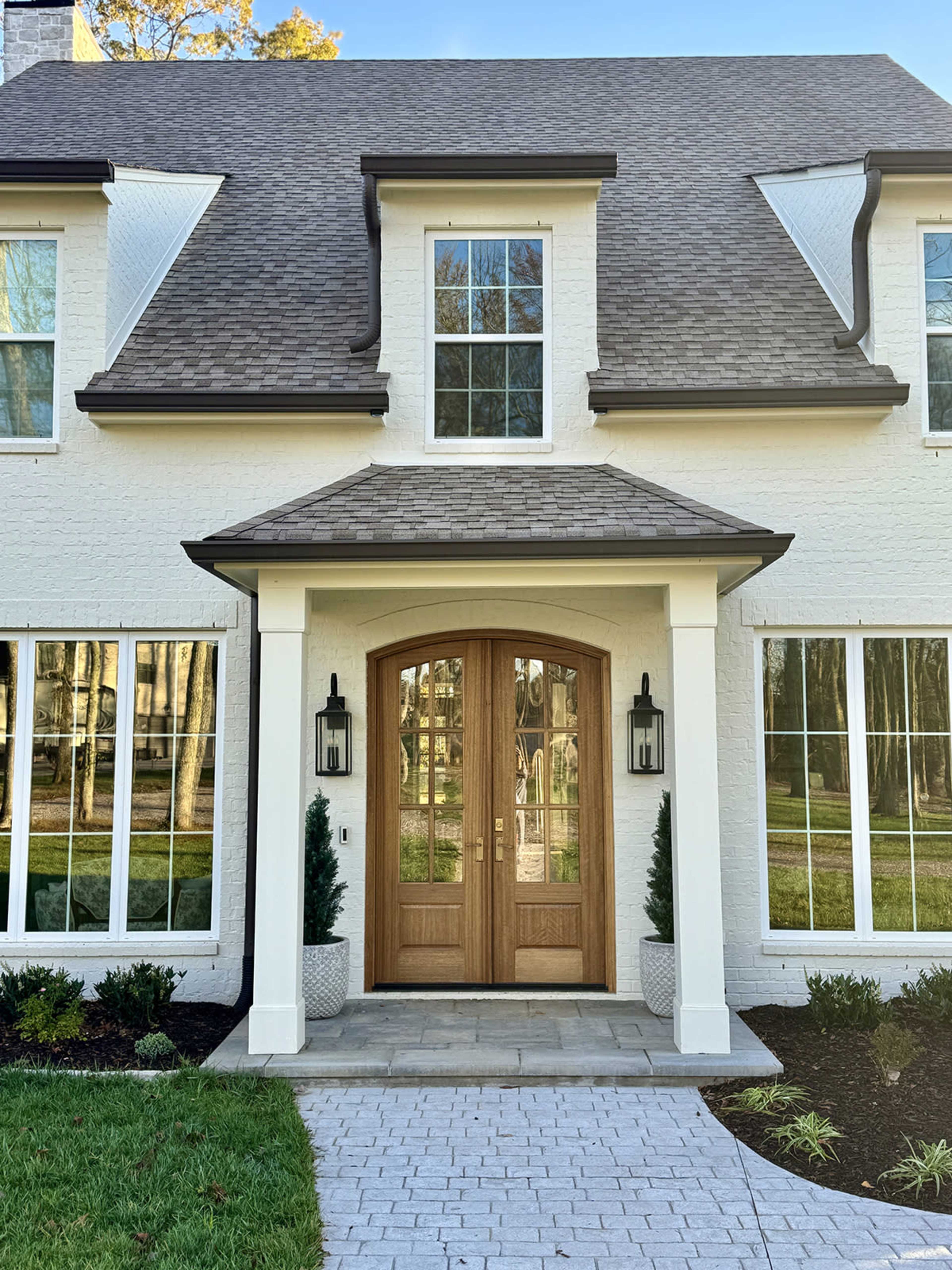 The image shows a two-story house with a symmetric façade, featuring a large wooden double door and two small trees in planters on either side of the entrance.