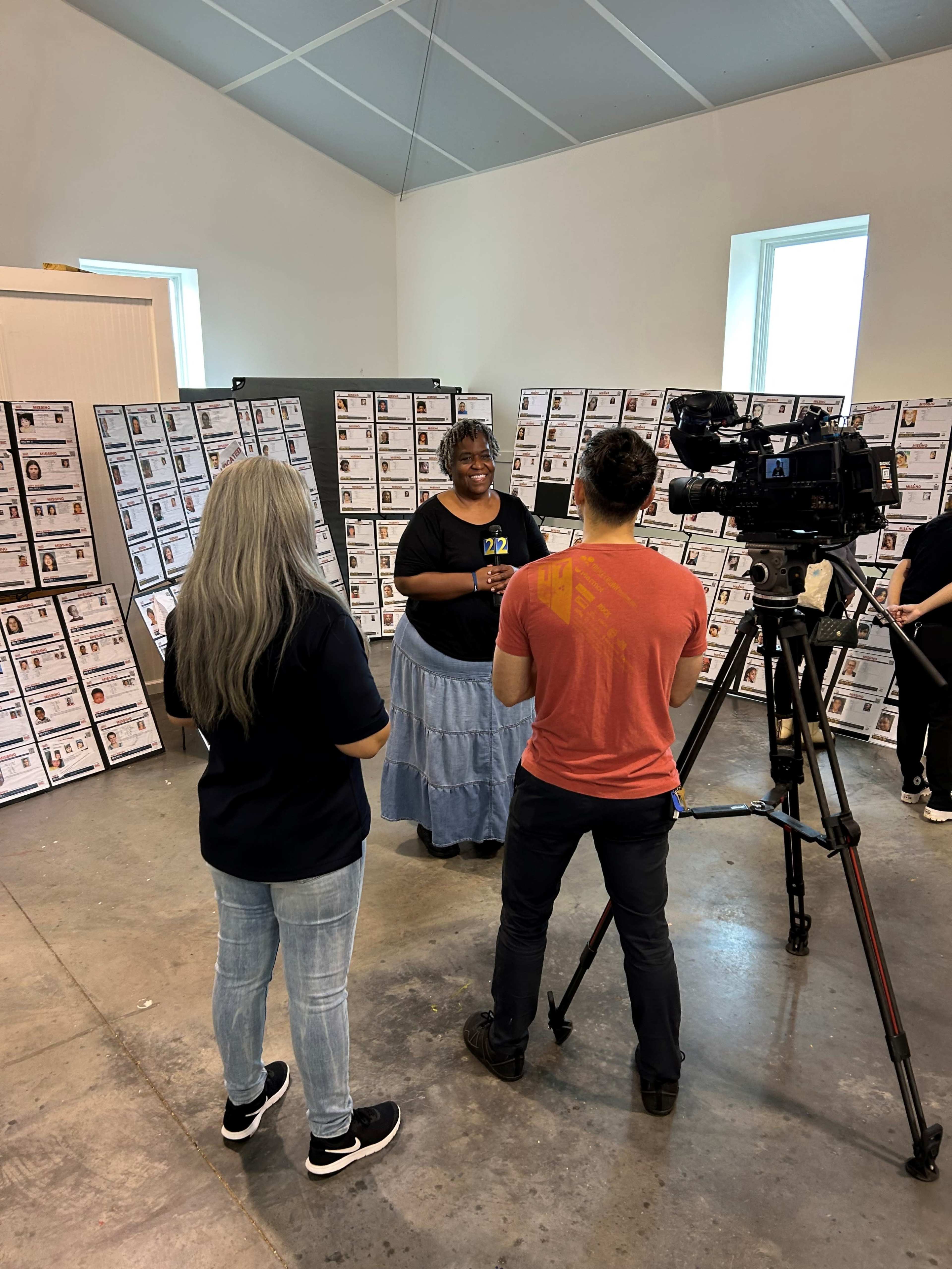 A woman in a blue skirt is being interviewed by a reporter and a camera operator in a room filled with posters on the walls.