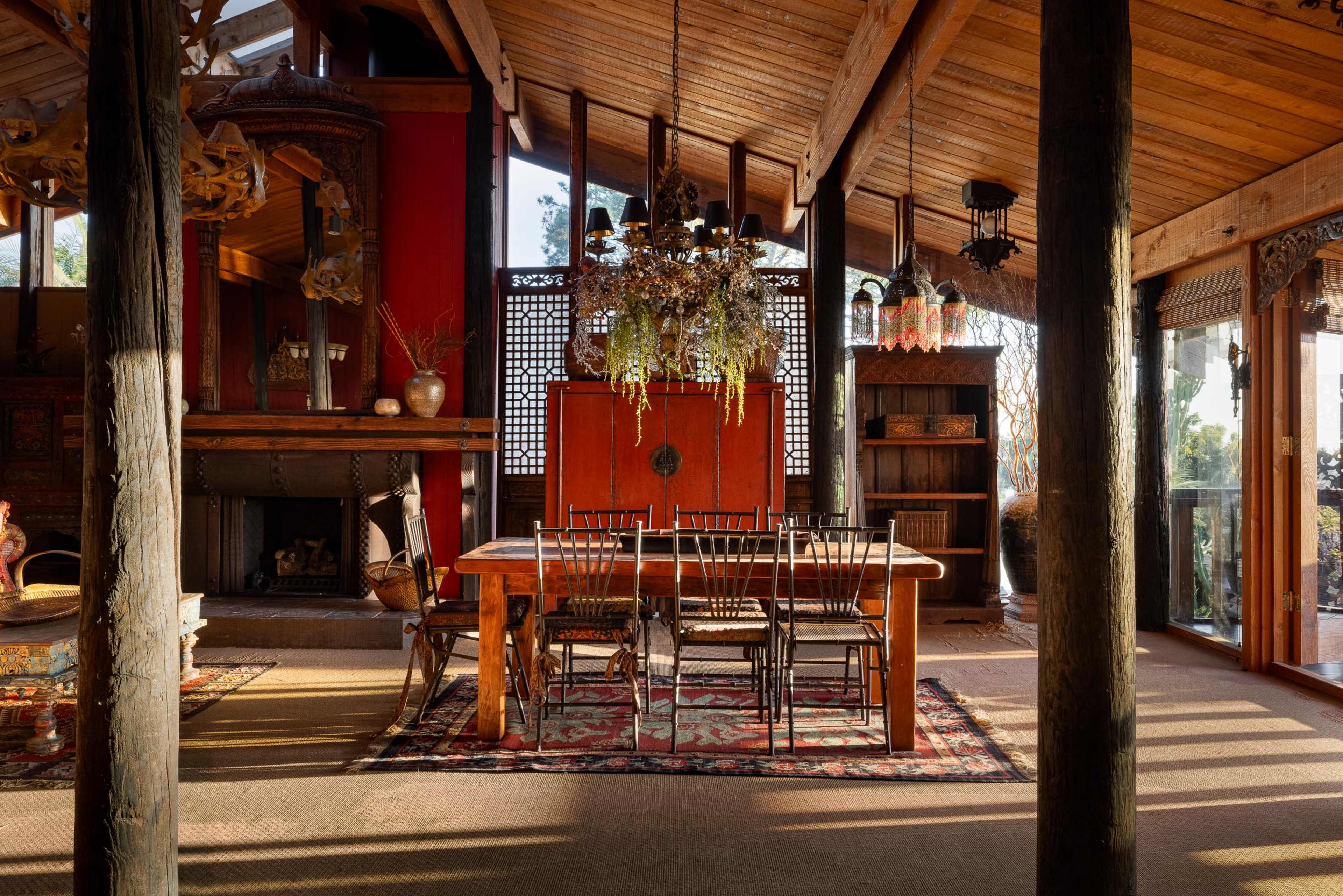 The image shows a spacious interior of a house featuring a wooden dining table surrounded by black chairs, set against a backdrop of large windows and rustic decor elements.
