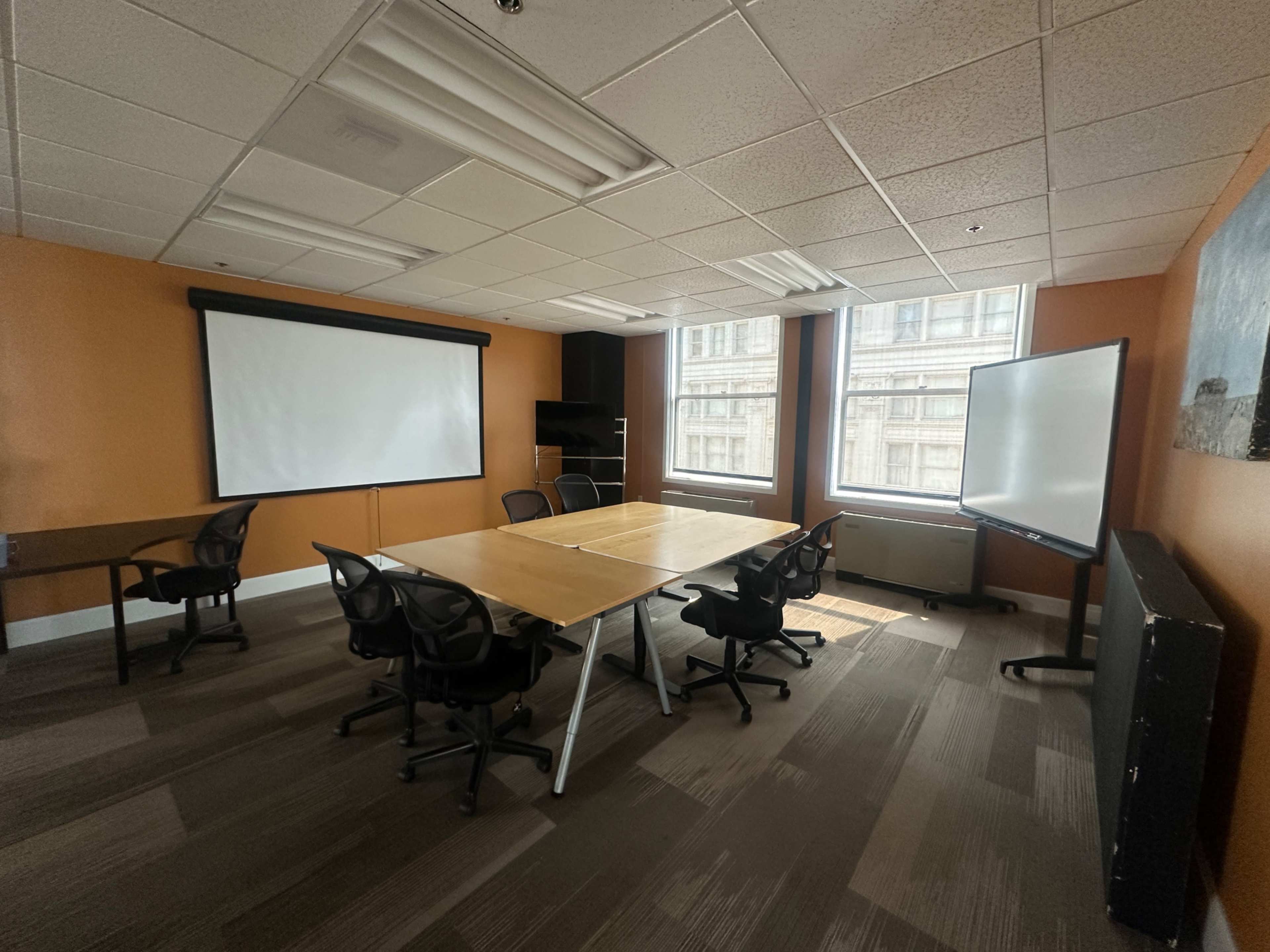 The image shows a vacant conference room with two large windows, a wooden table surrounded by black chairs, and two projector screens on opposite walls.