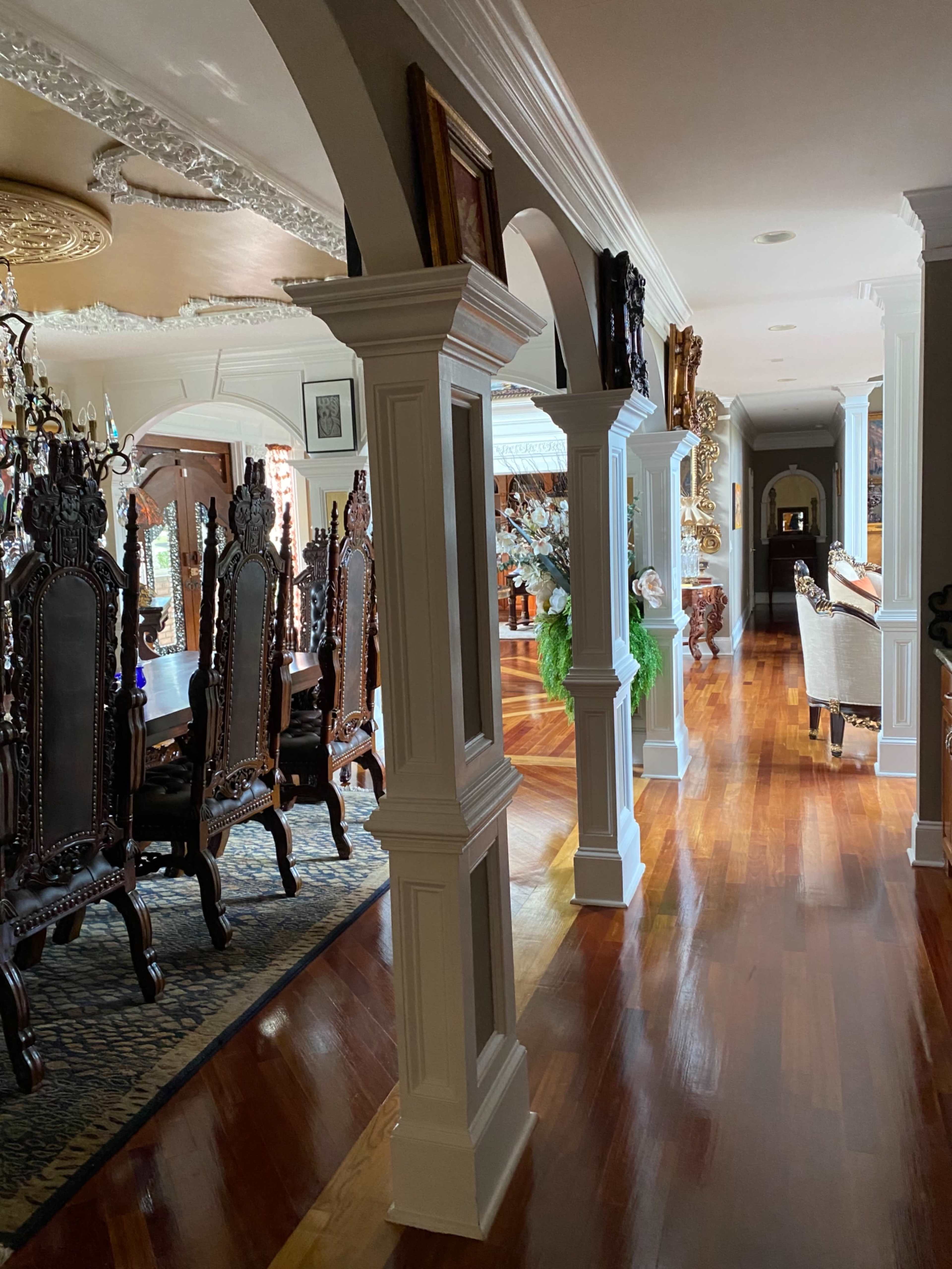 A long hallway with wooden floors, featuring ornate columns and leading to a dining area with a large table and intricately designed chairs.