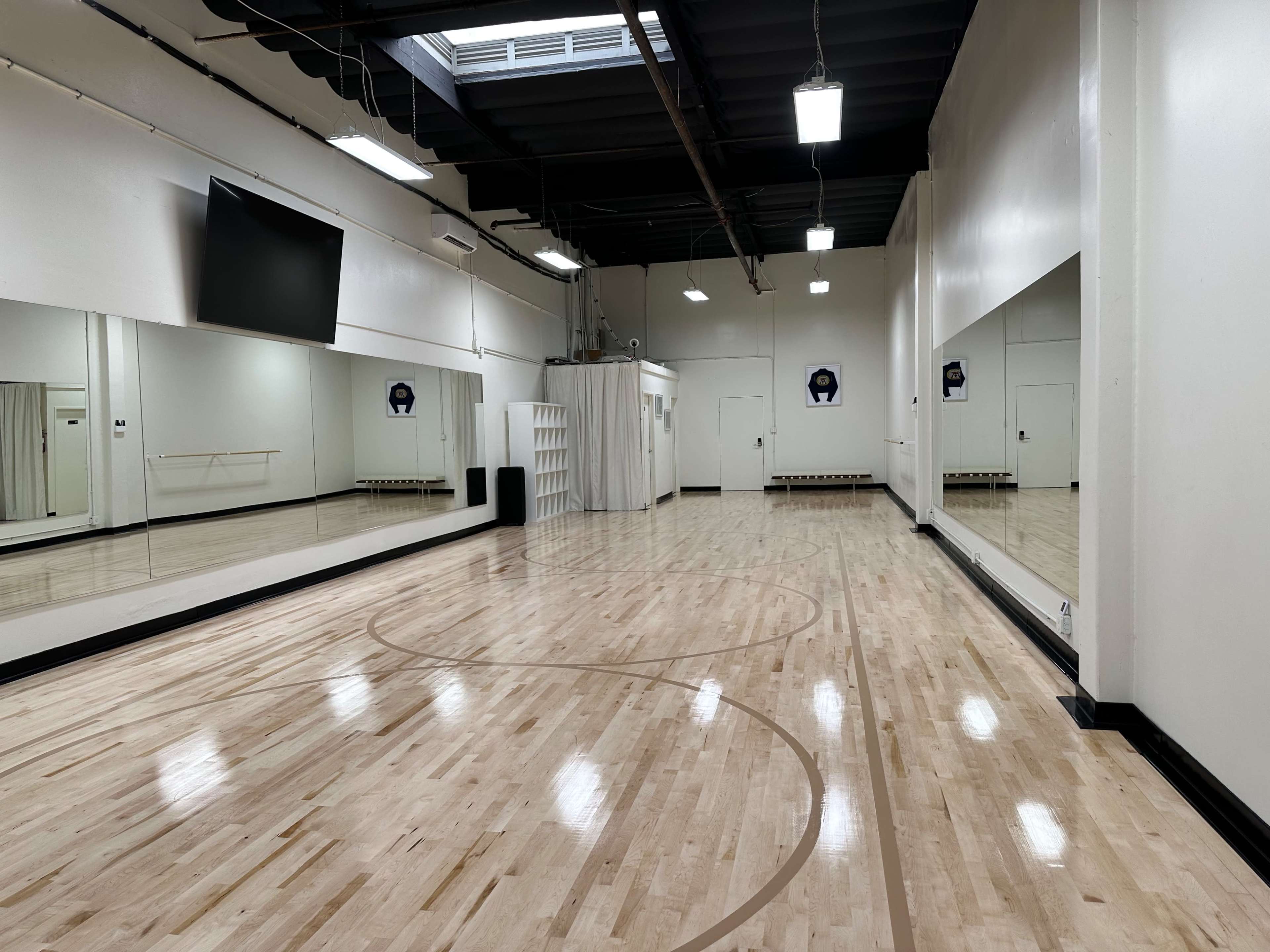 The image shows a spacious dance studio with wooden floors, large mirrors on the walls, and a mounted television.