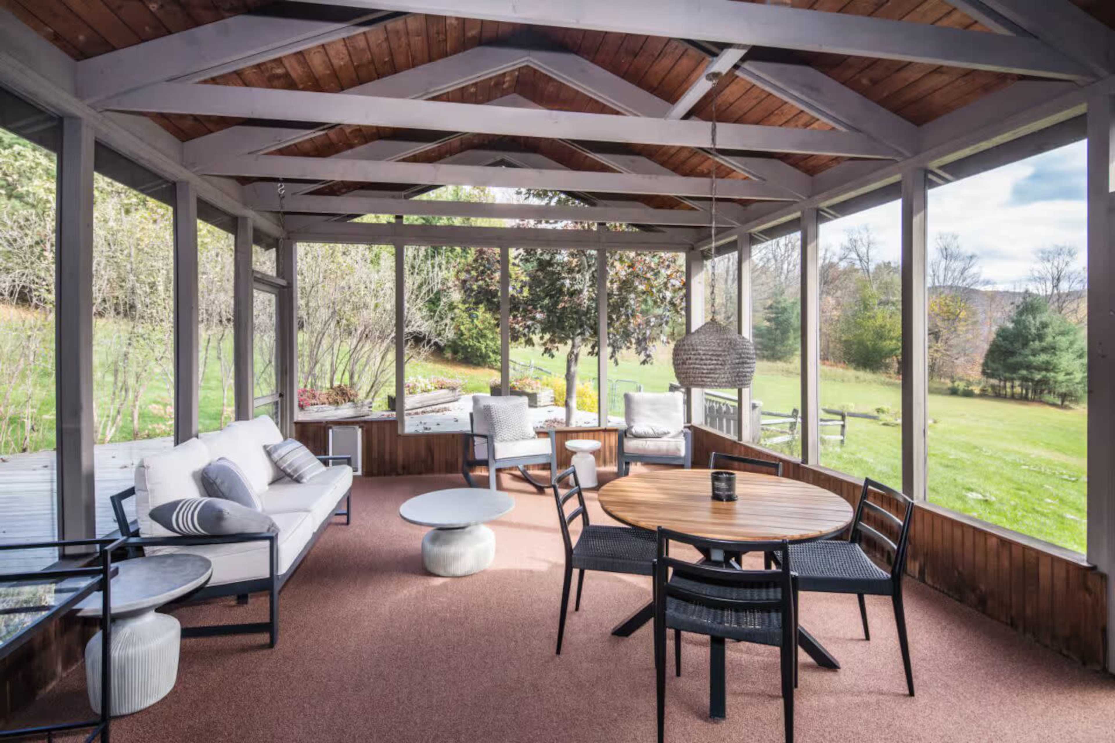 The image shows a screened-in porch with a seating area, a round dining table, and views of a grassy landscape beyond.