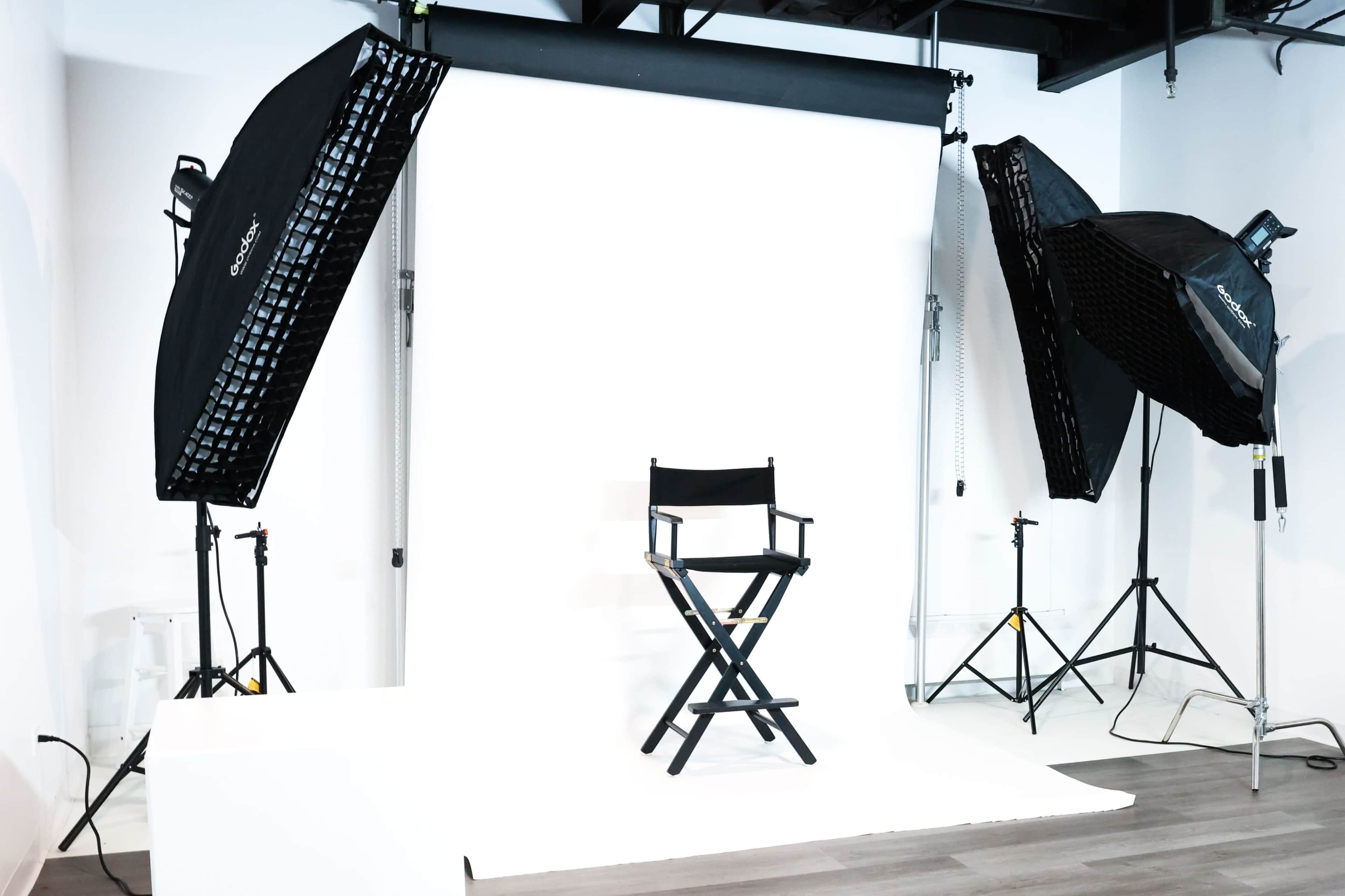 The image shows a photography studio setup with a black director's chair positioned in front of a plain white backdrop, flanked by two large softbox lights.