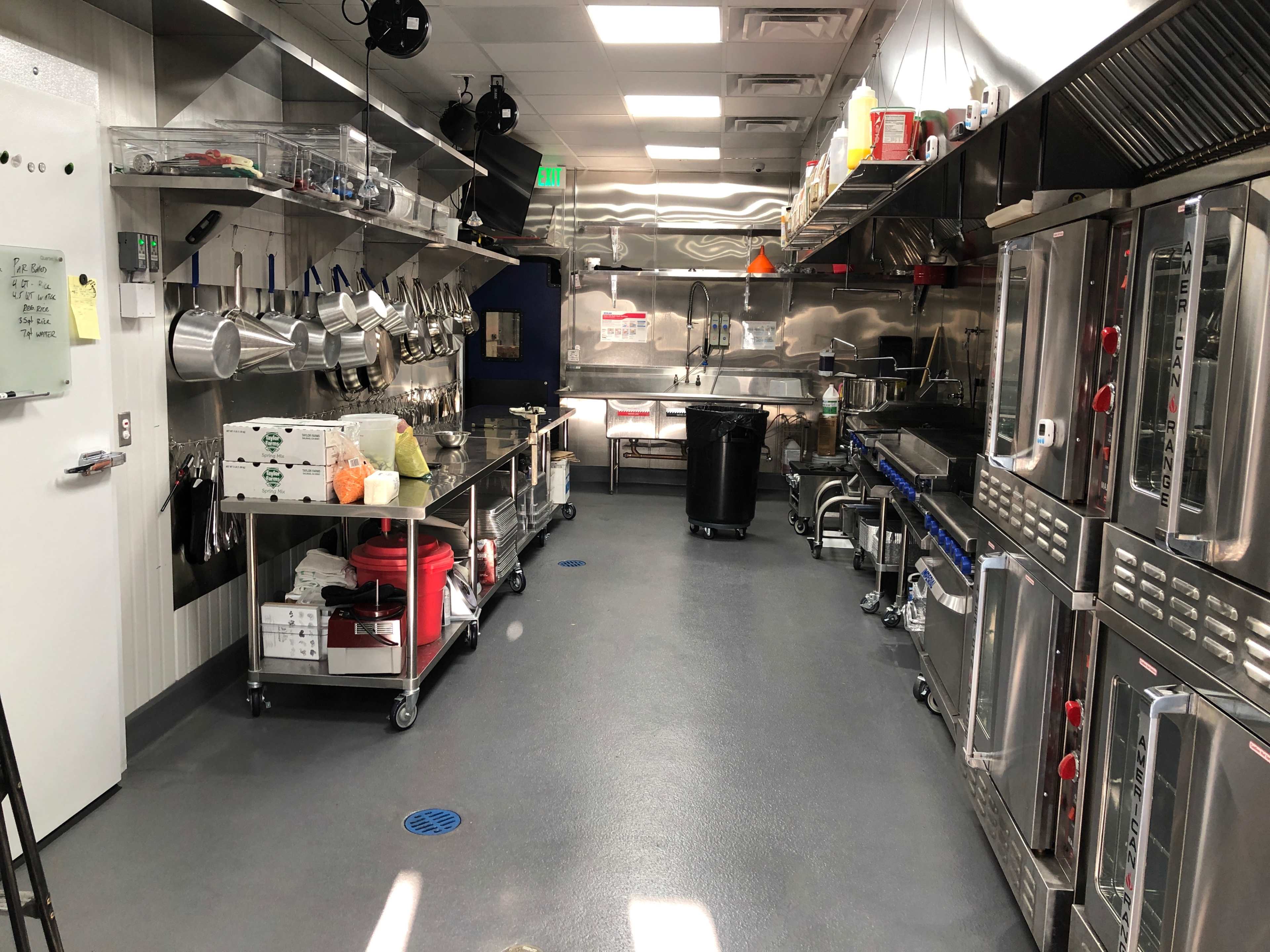 The image shows a modern commercial kitchen equipped with stainless steel appliances, storage shelves, and cooking utensils arranged neatly.