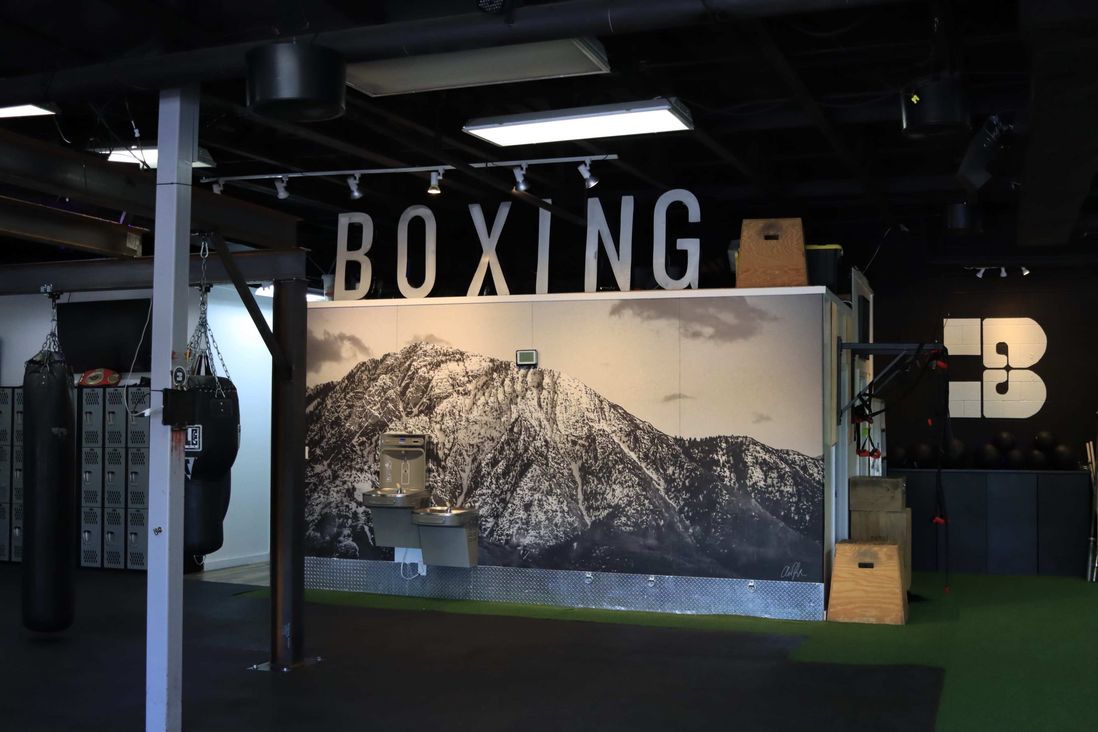 The image shows a boxing gym interior featuring a large mountain mural, a drinking fountain, and heavy bags hanging from the ceiling.