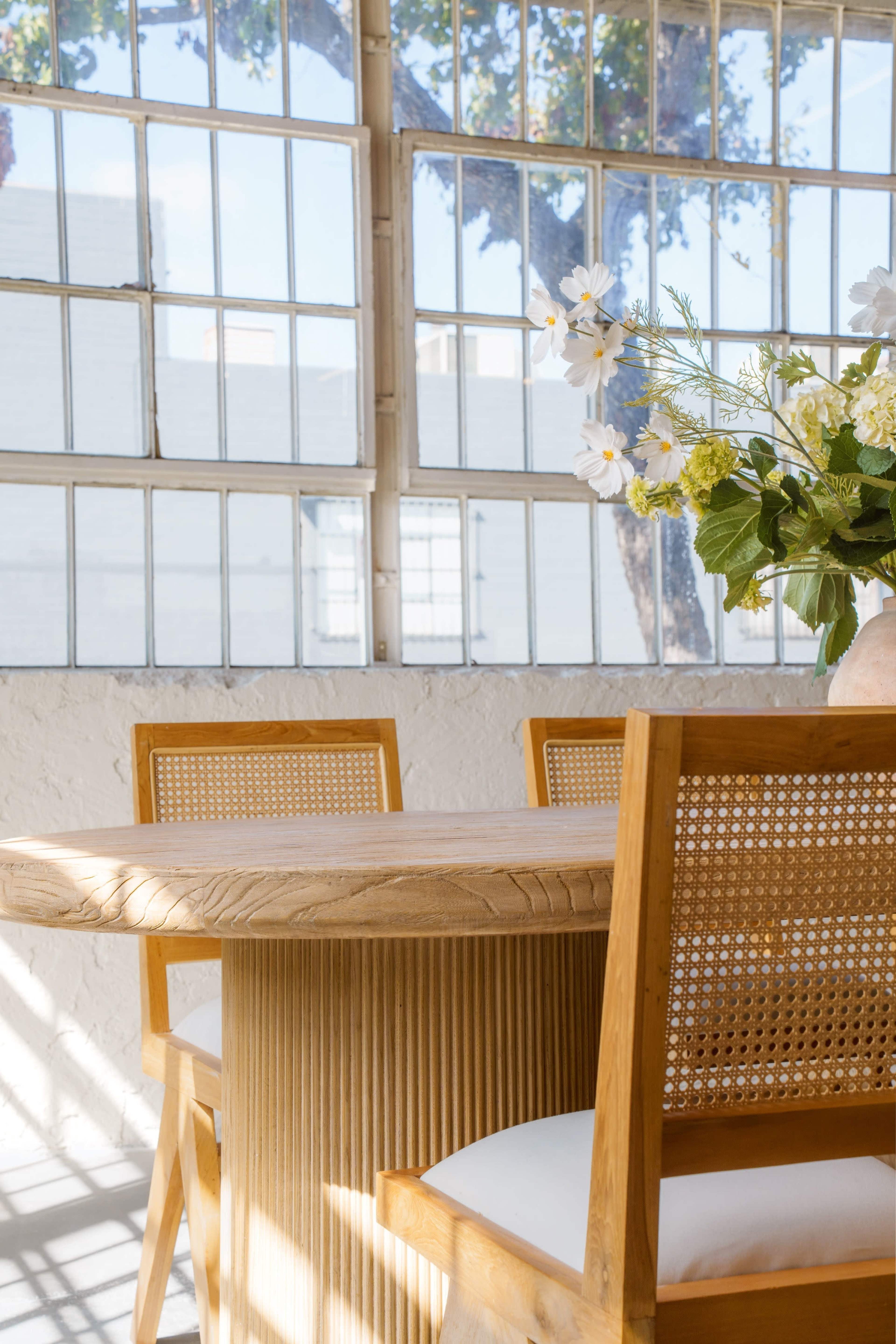A round wooden table with two caned chairs is positioned near large windows adorned with natural light, along with a vase of flowers on the table.