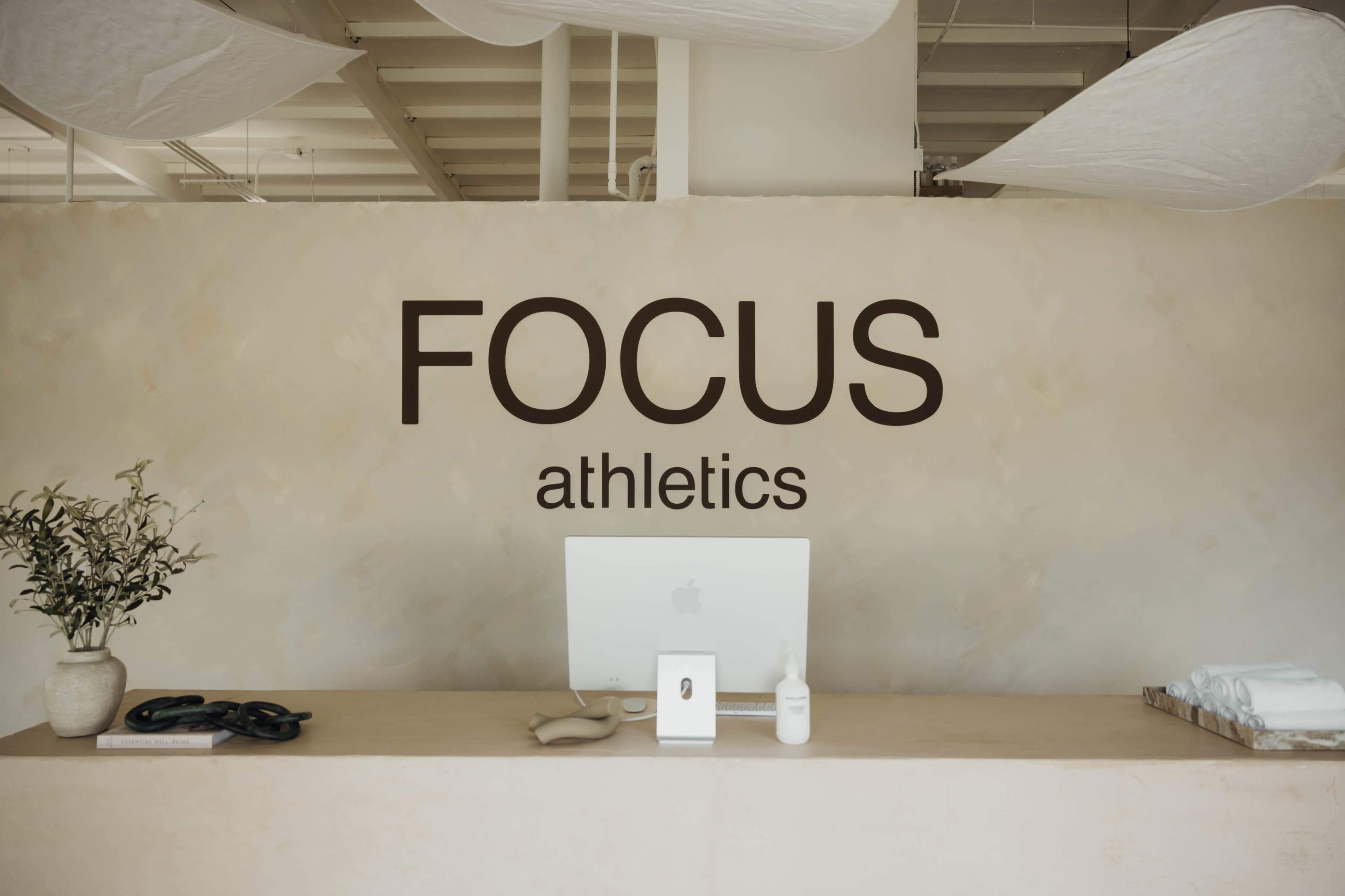 A minimalistic reception area features a desk with an iMac, a plant, and towels, all set against a wall displaying the words "FOCUS athletics."