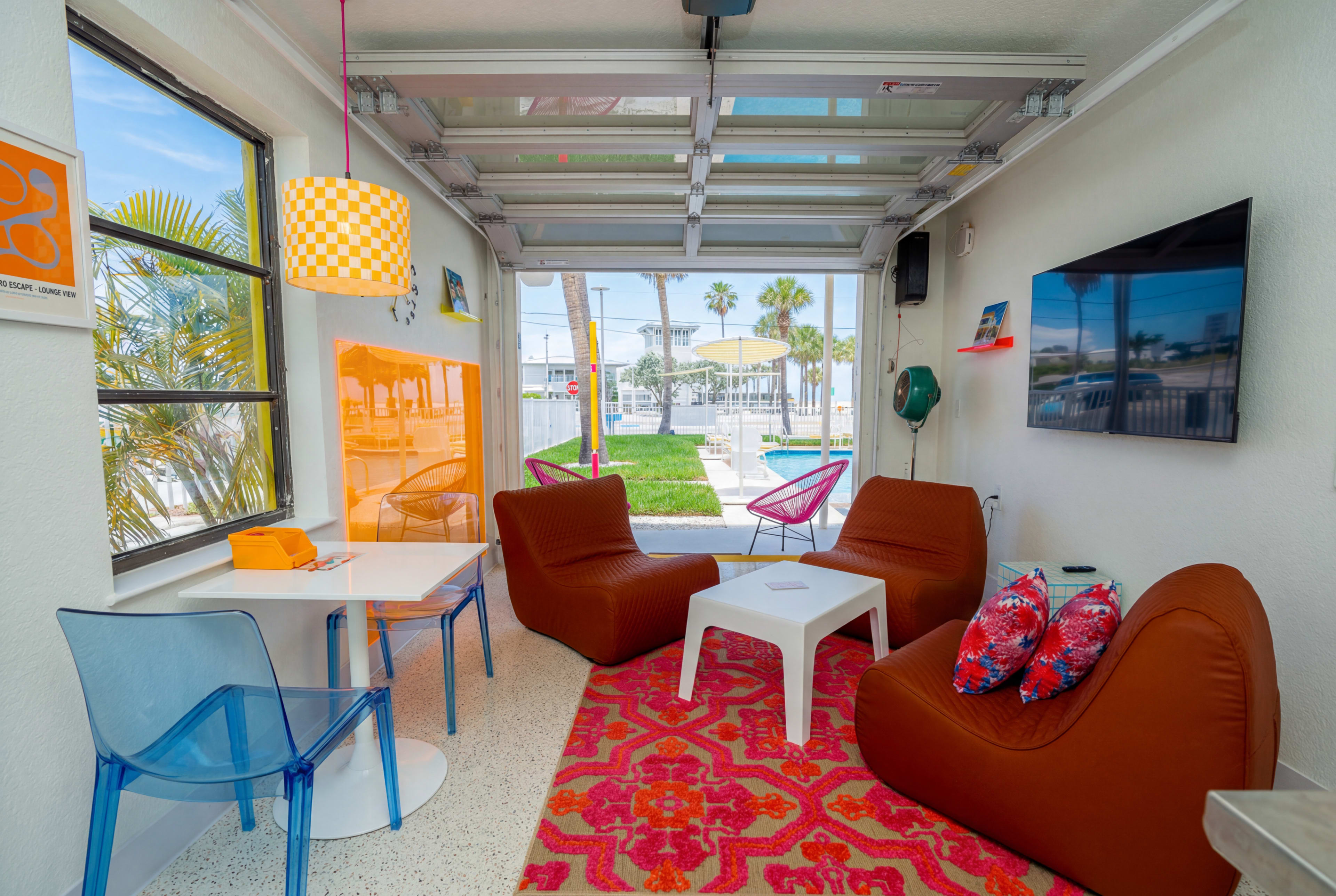 The image shows a modern, vibrant living space with colorful furniture, a large window, and a garage door that opens to an outdoor area with a lawn and swimming pool.