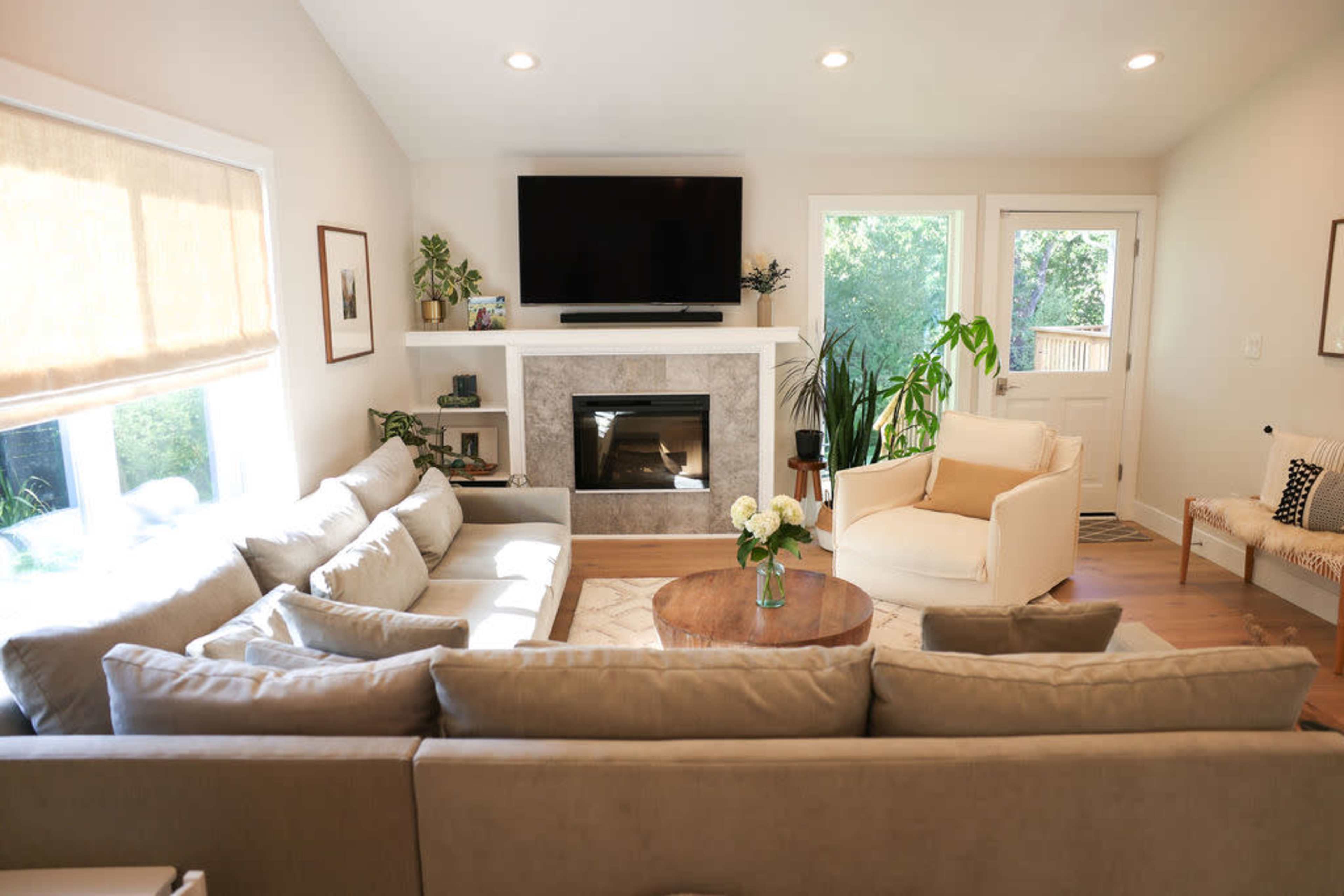 A modern living room with a large sectional sofa, a circular coffee table, a fireplace with a flat-screen TV above it, and large windows letting in natural light.