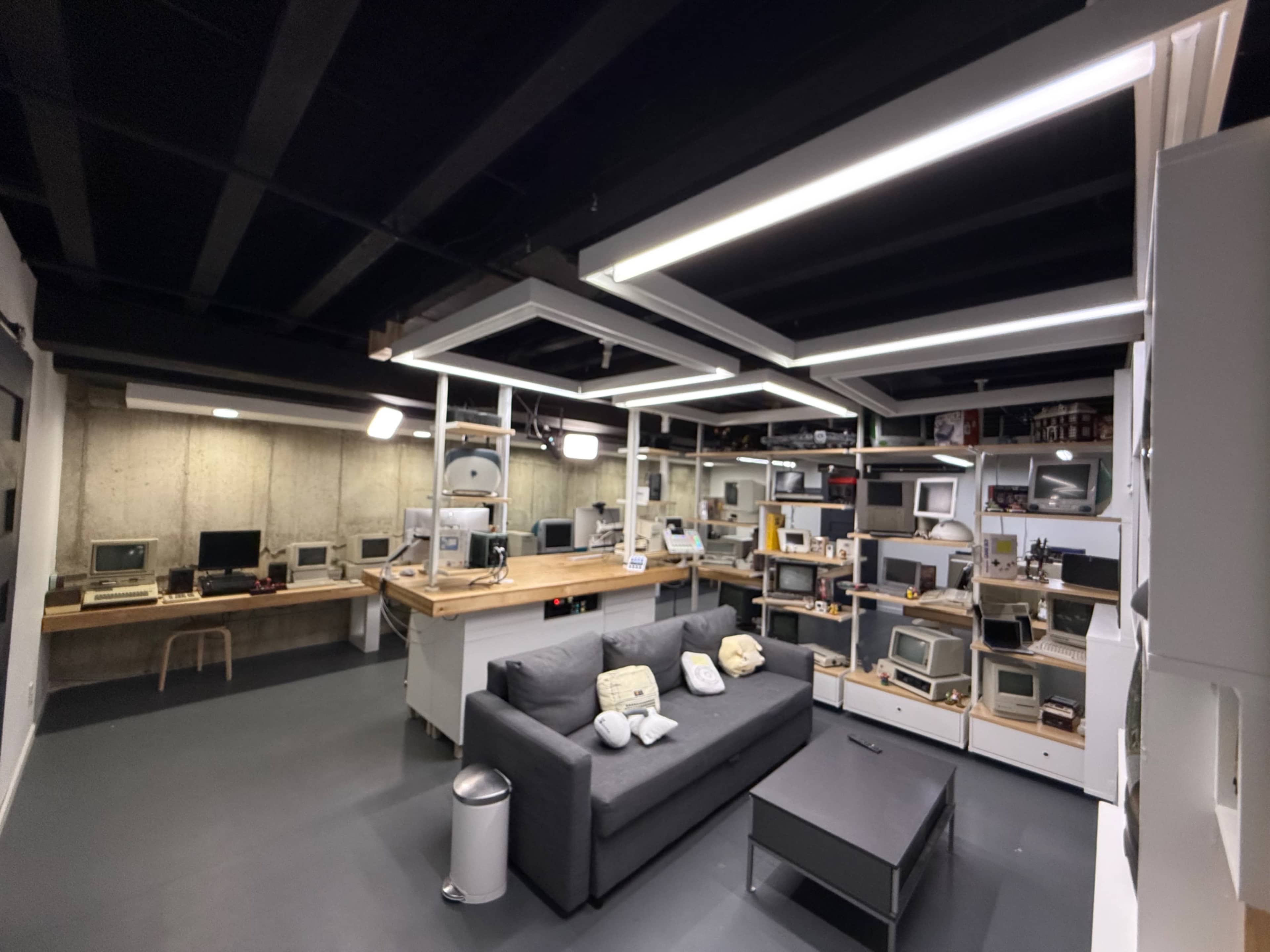 The image shows a modern, minimalist lounge area in a basement, featuring a gray sofa, a coffee table, and several vintage computers displayed on shelves.