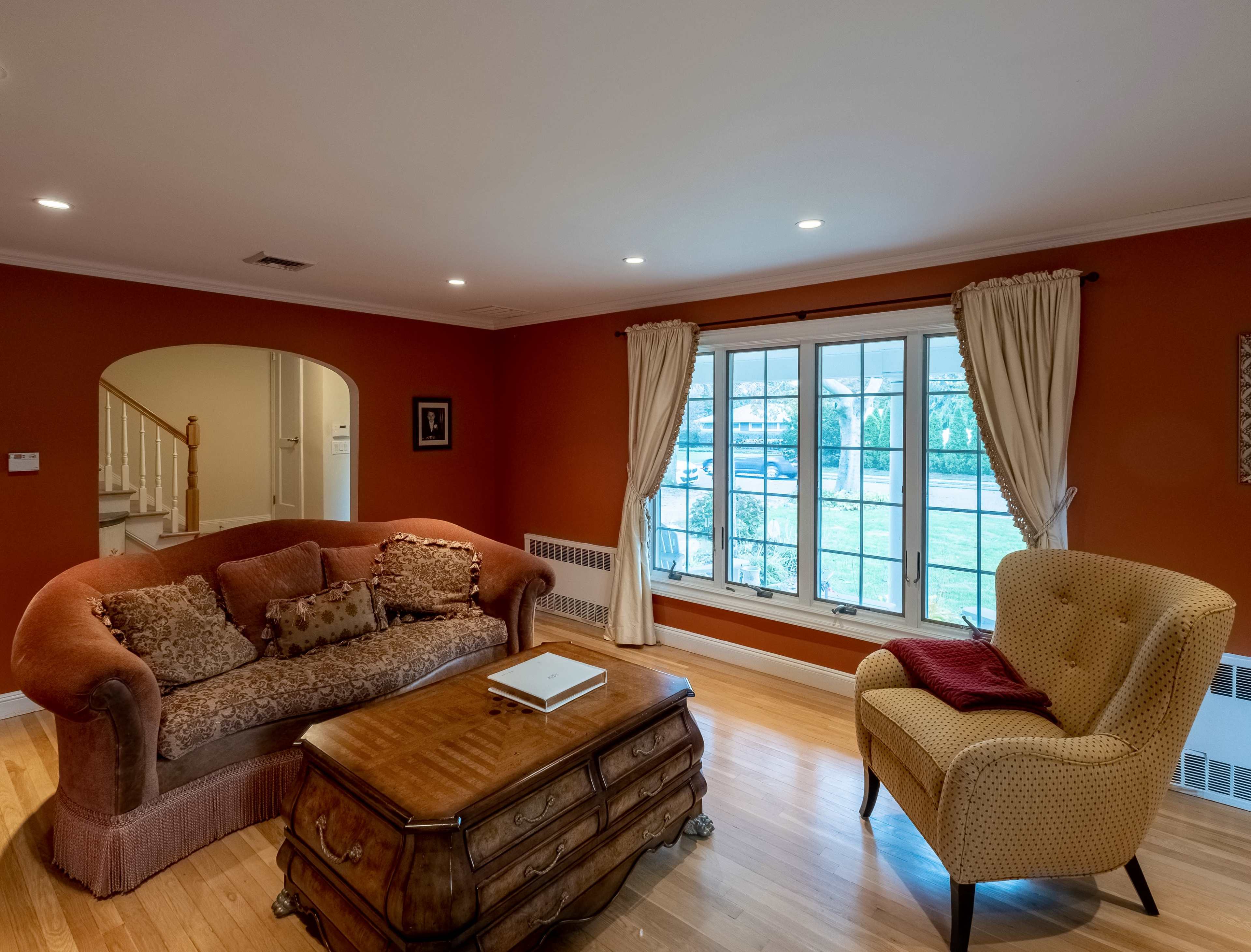 The living room features a brown sofa and a yellow armchair, a wooden coffee table, hardwood flooring, and a large window with curtains that overlooks a garden.