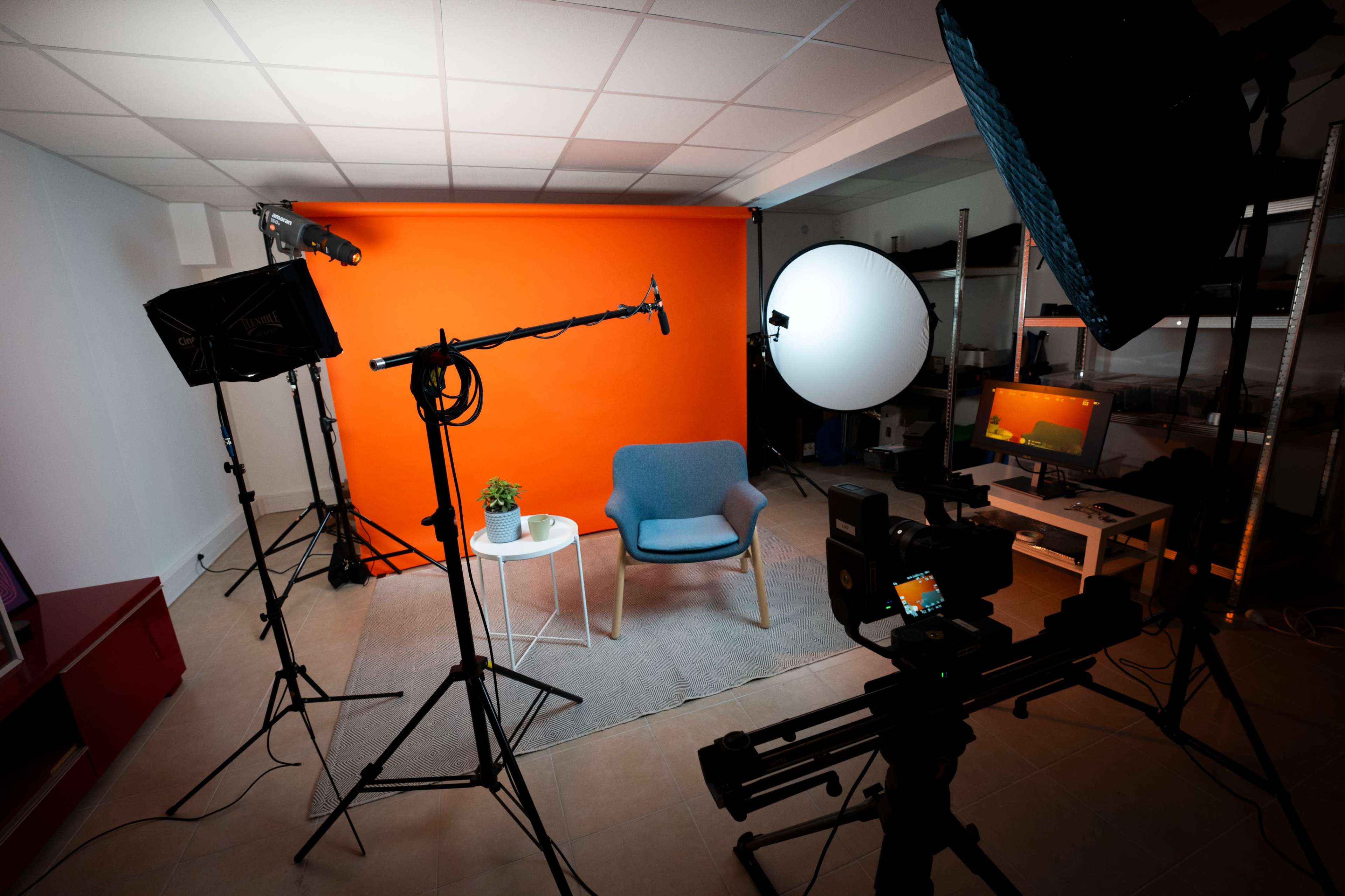 A well-lit photography studio features a blue chair and a small table against a bright orange backdrop, surrounded by multiple lights and cameras.