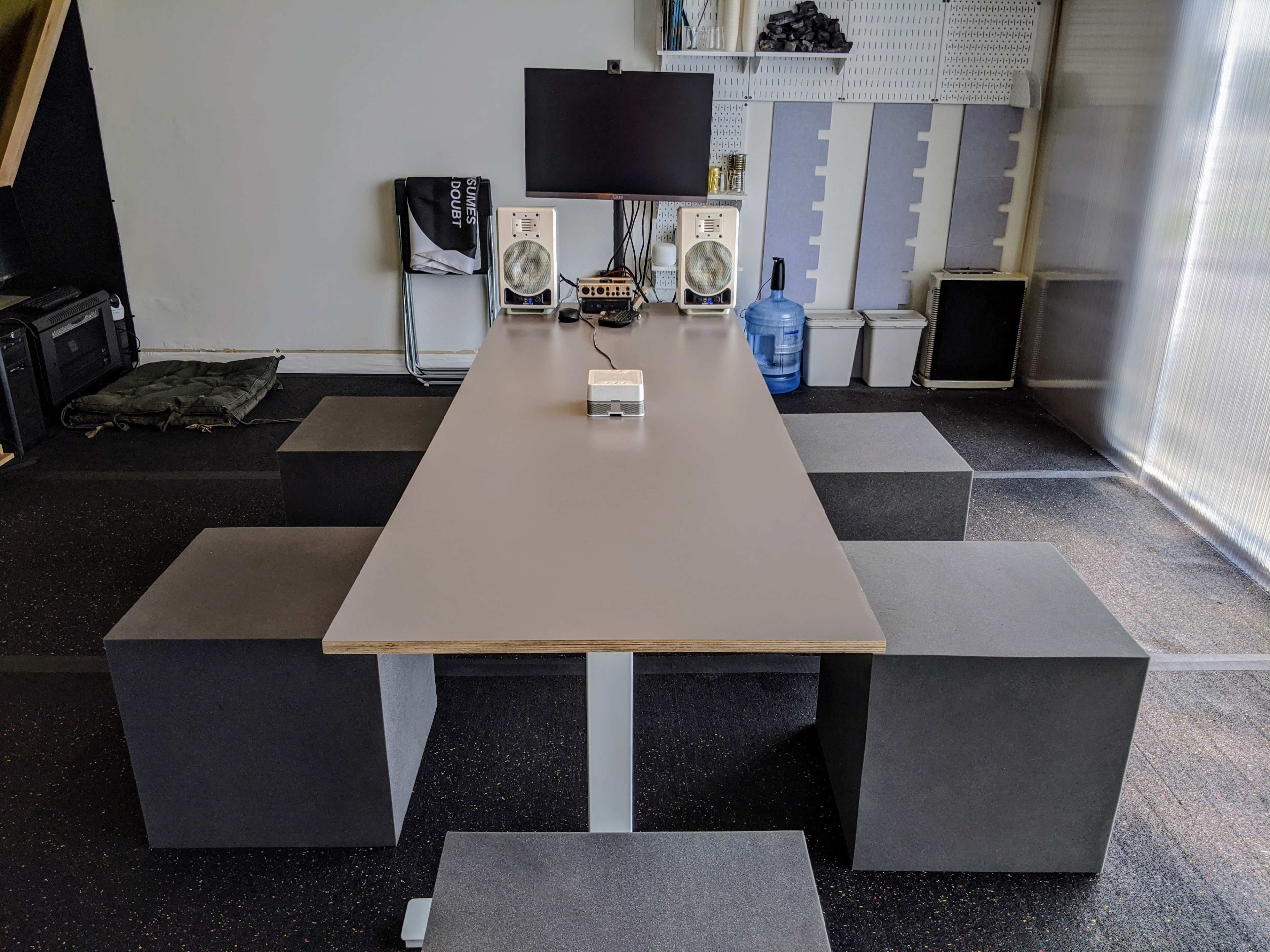 A long gray table is set up in a room with four gray cube seats, flanked by a computer monitor and speakers.