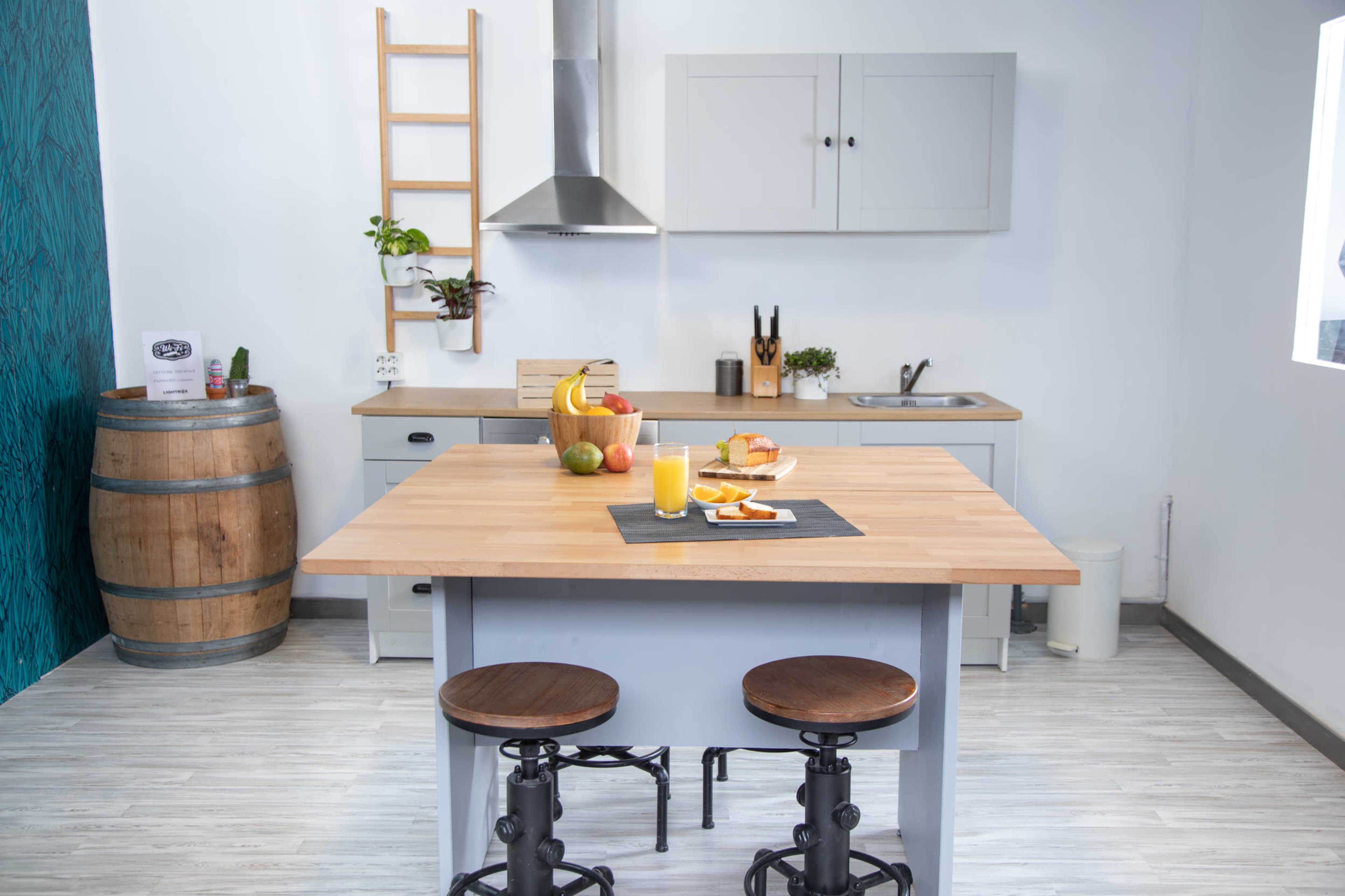 A modern kitchen features a wooden island with a variety of fruits and a drink, accompanied by two stools, and cabinets lined against a wall.