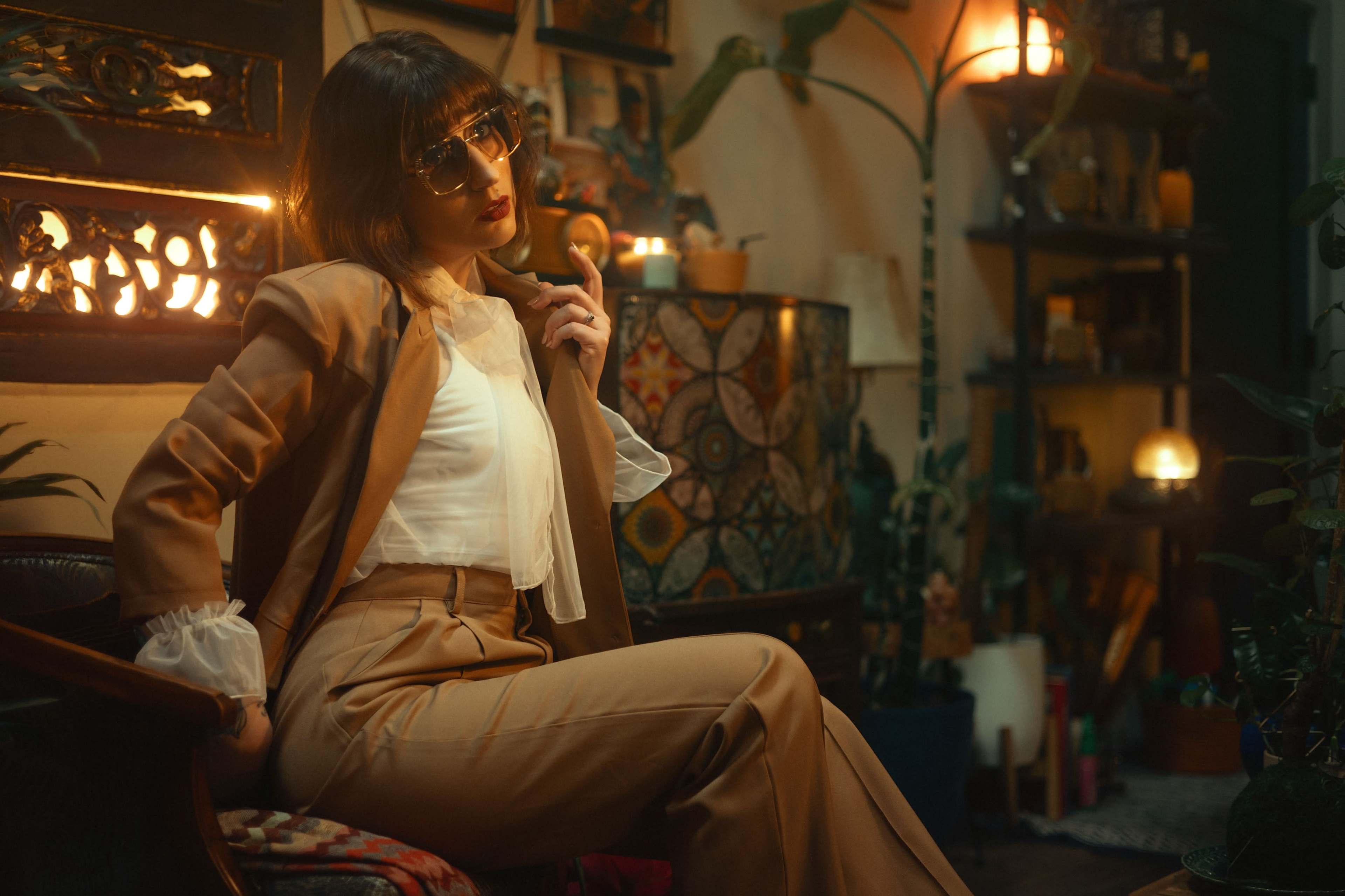 A person in a tan suit with a white blouse sits on a chair in a warmly lit room filled with plants and decorative items.