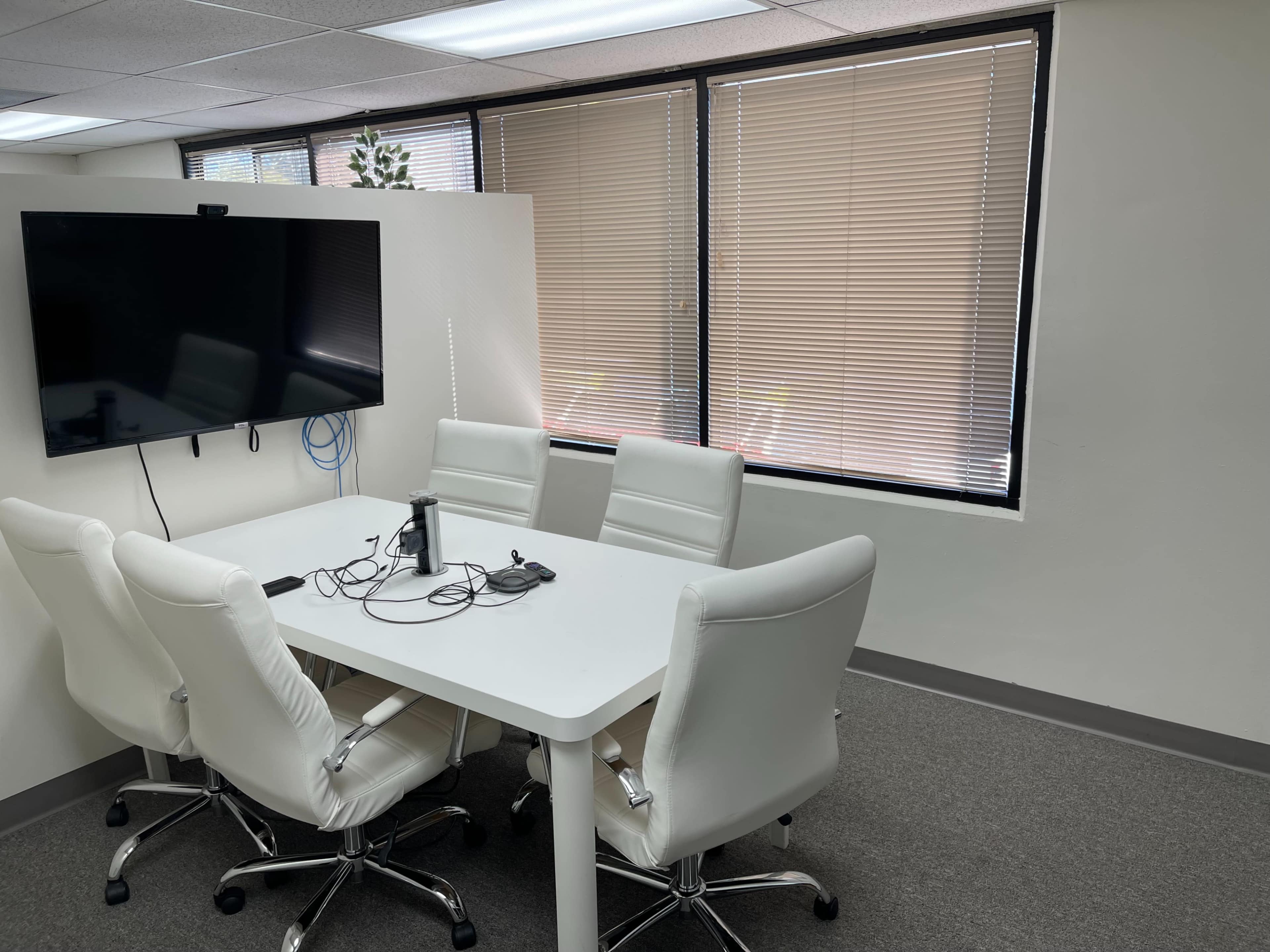 A conference room features a white table surrounded by four white chairs, with a large screen mounted on the wall and natural light coming through window blinds.