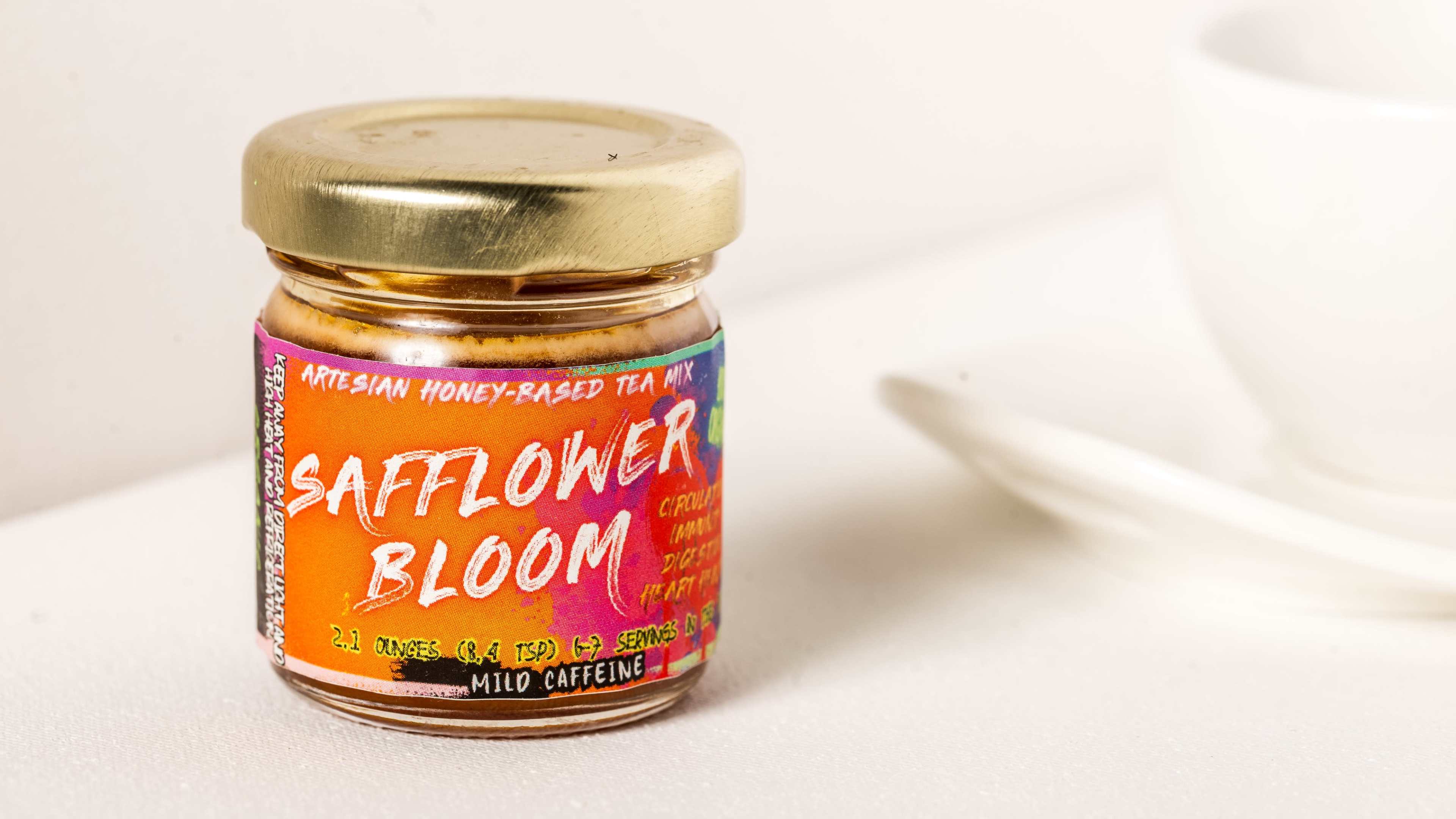 A small glass jar labeled "Safflower Bloom" contains artisan honey-based tea and is placed near a white cup.