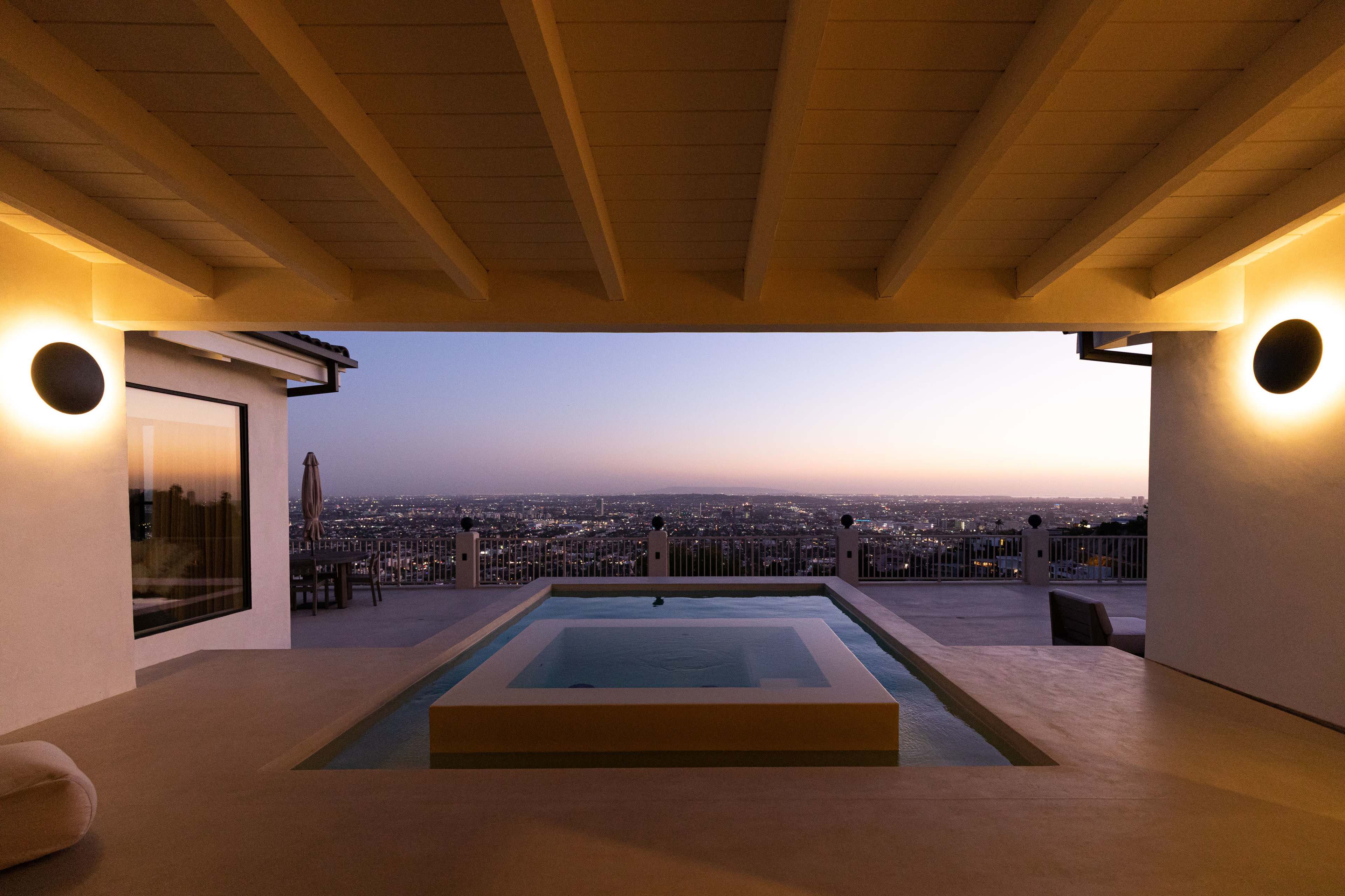 A modern outdoor space with a pool overlooking a cityscape at dusk, framed by a spacious overhang.