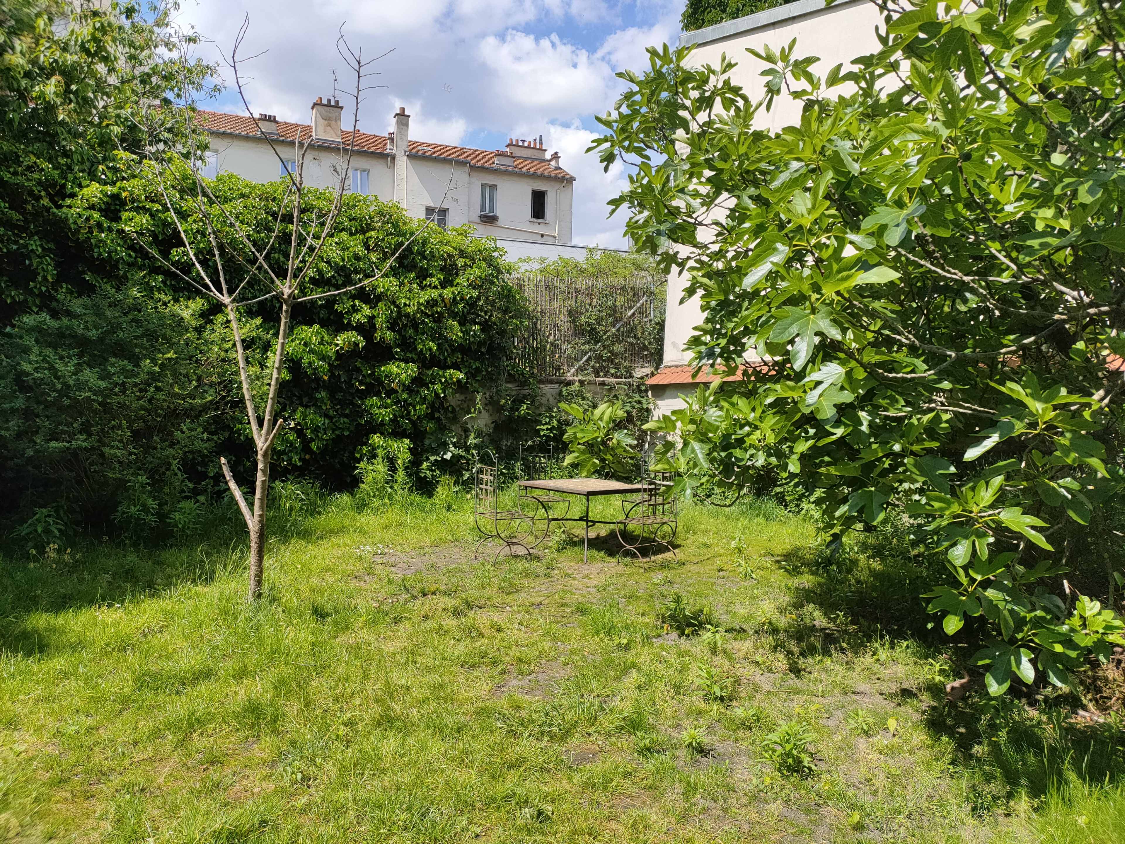 The image shows a grassy garden with two trees and a round metal table surrounded by greenery, set against a backdrop of buildings.
