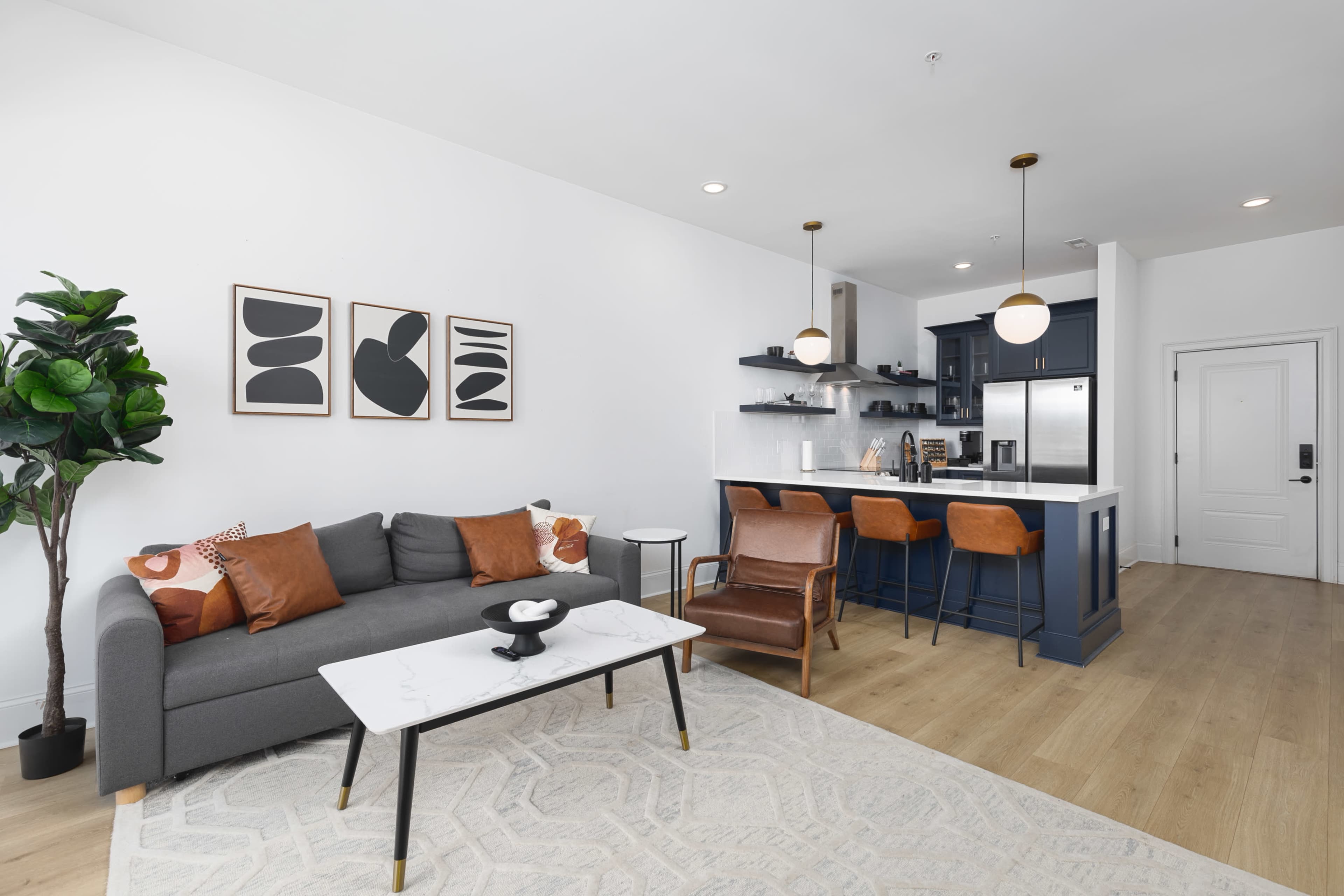 A modern living room features a gray sofa with brown cushions, a patterned area rug, a marble coffee table, and an open kitchen with dark cabinets and pendant lighting.