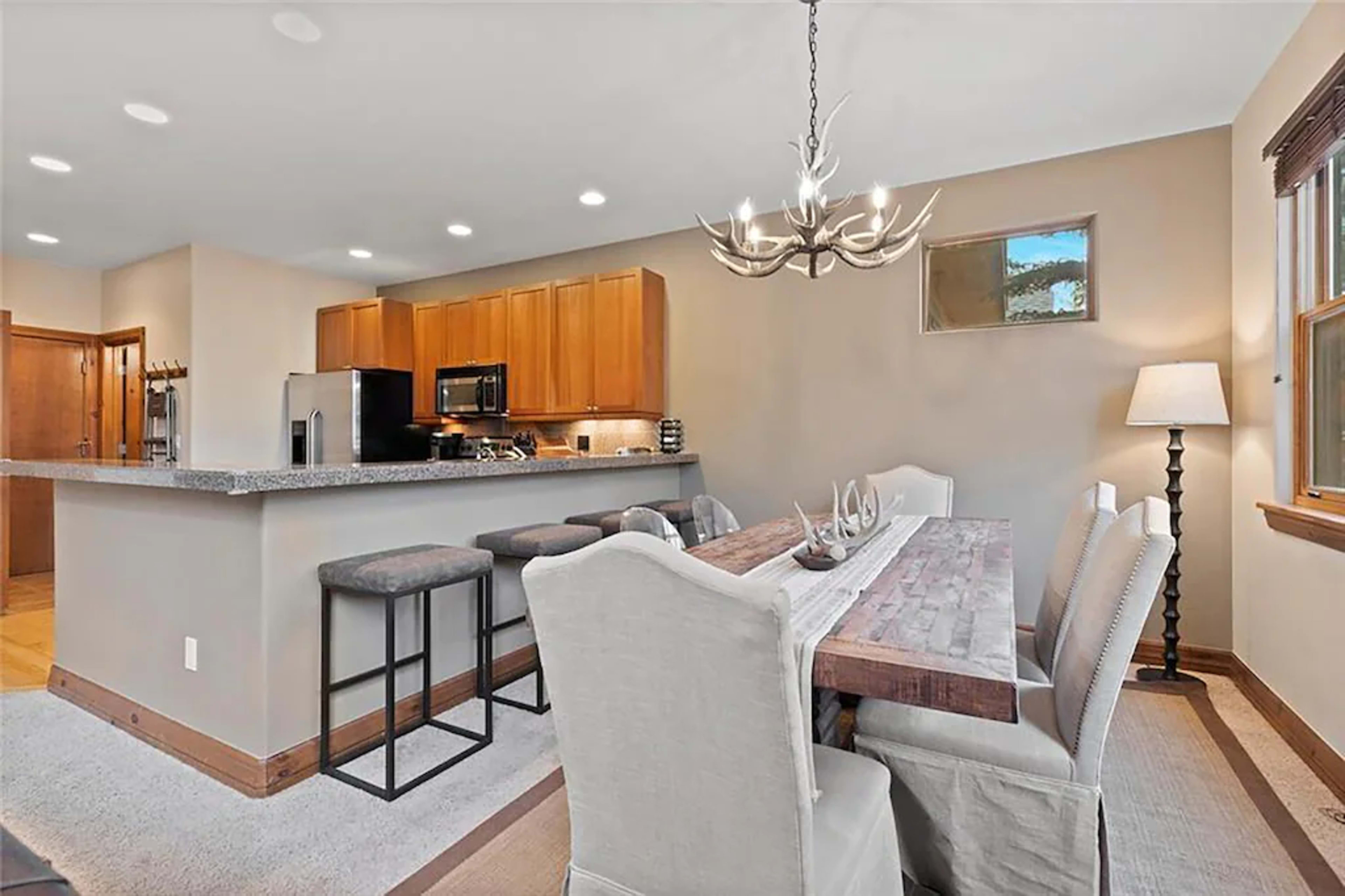 A modern kitchen and dining area with a wooden dining table and six upholstered chairs, adjacent to a kitchen featuring wooden cabinetry and bar seating.