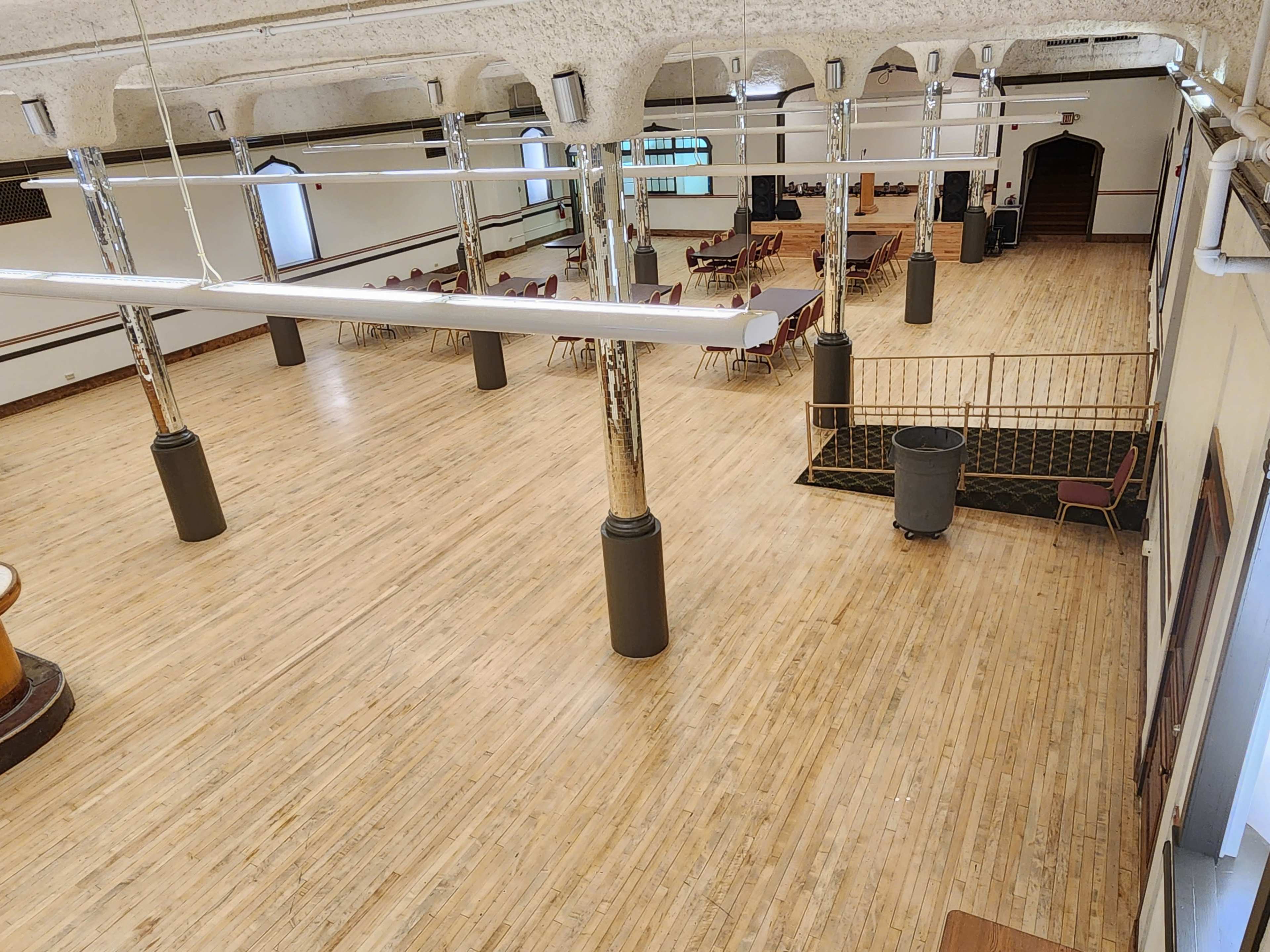 The image depicts a spacious interior with wooden flooring, several columns, and tables arranged for an event in a well-lit hall.
