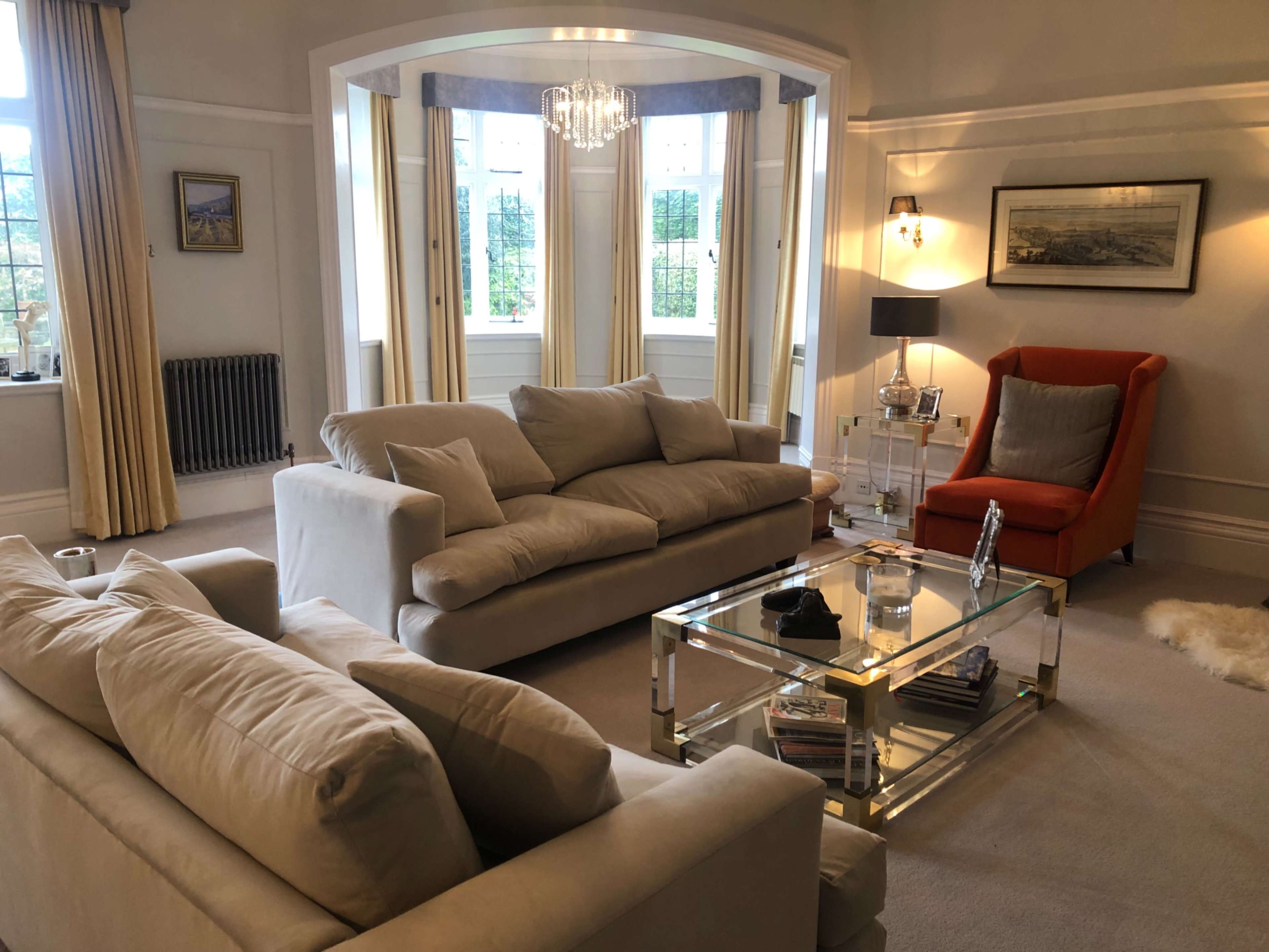 The living room features two beige sofas, an orange armchair, a glass coffee table, and large windows with sheer curtains.