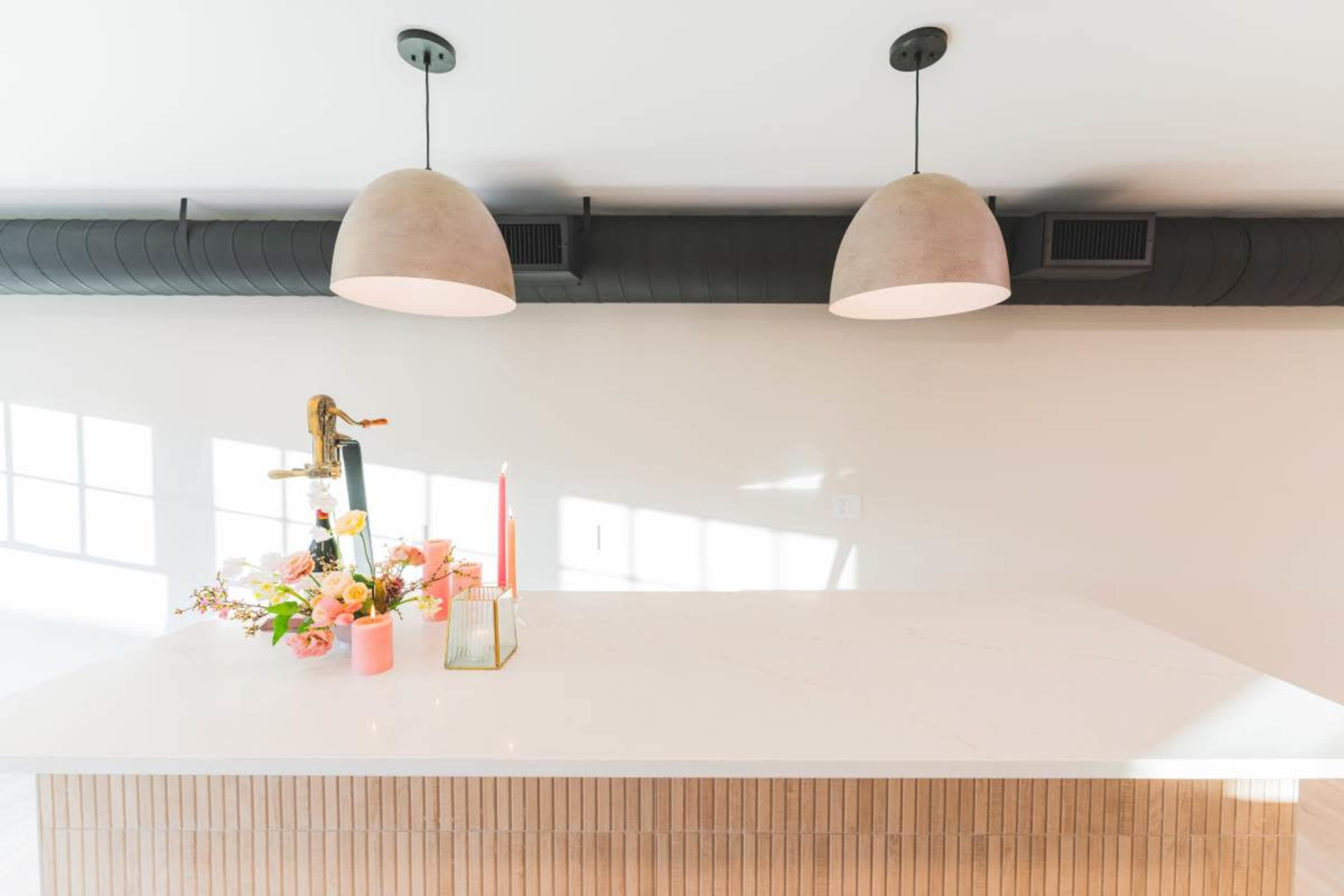 A modern kitchen features a white countertop with a floral arrangement and candles, beneath two pendant lights and a dark ventilation duct.