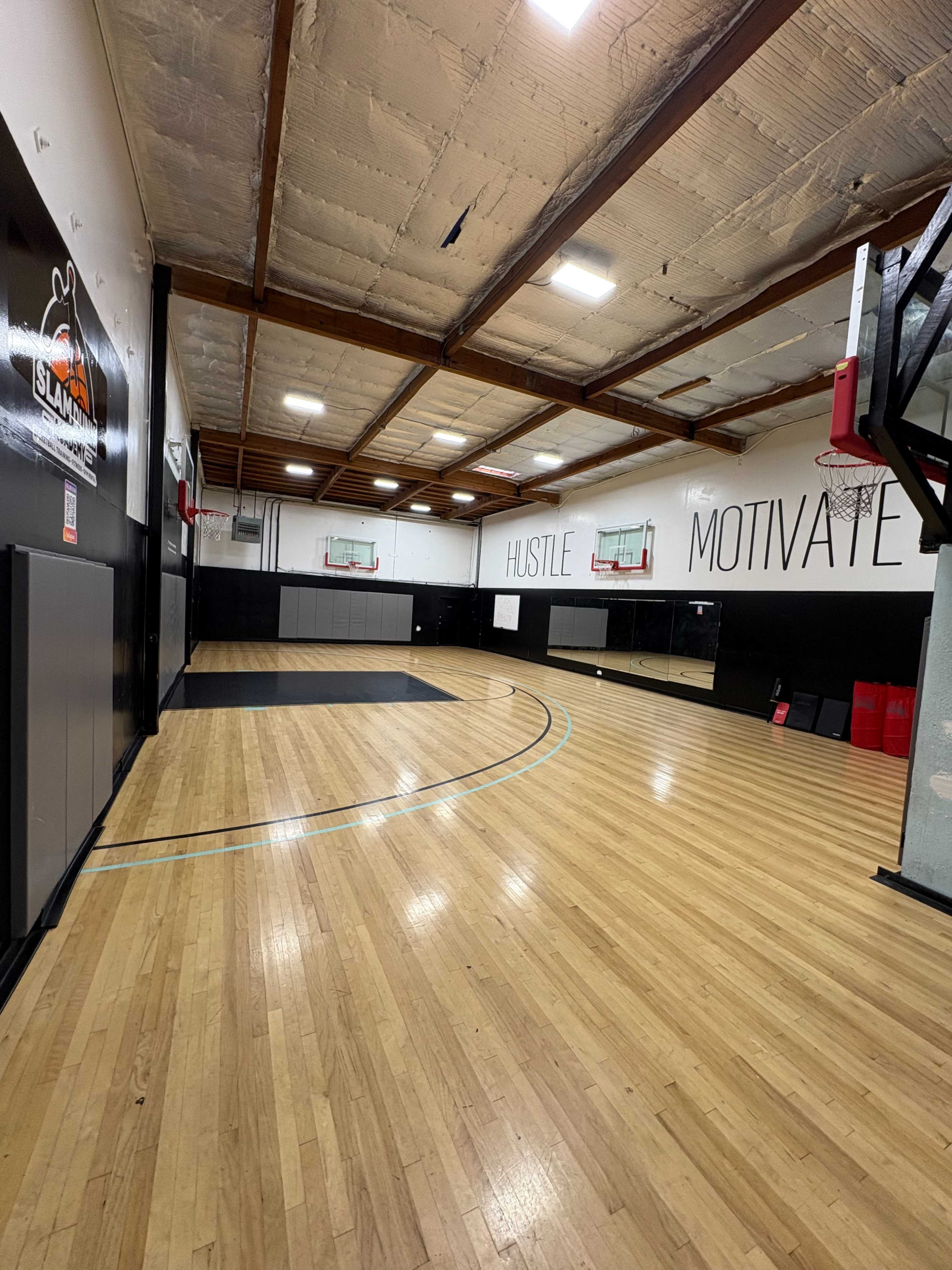 The image shows an indoor basketball court with wooden flooring, two basketball hoops, and motivational phrases painted on the walls.