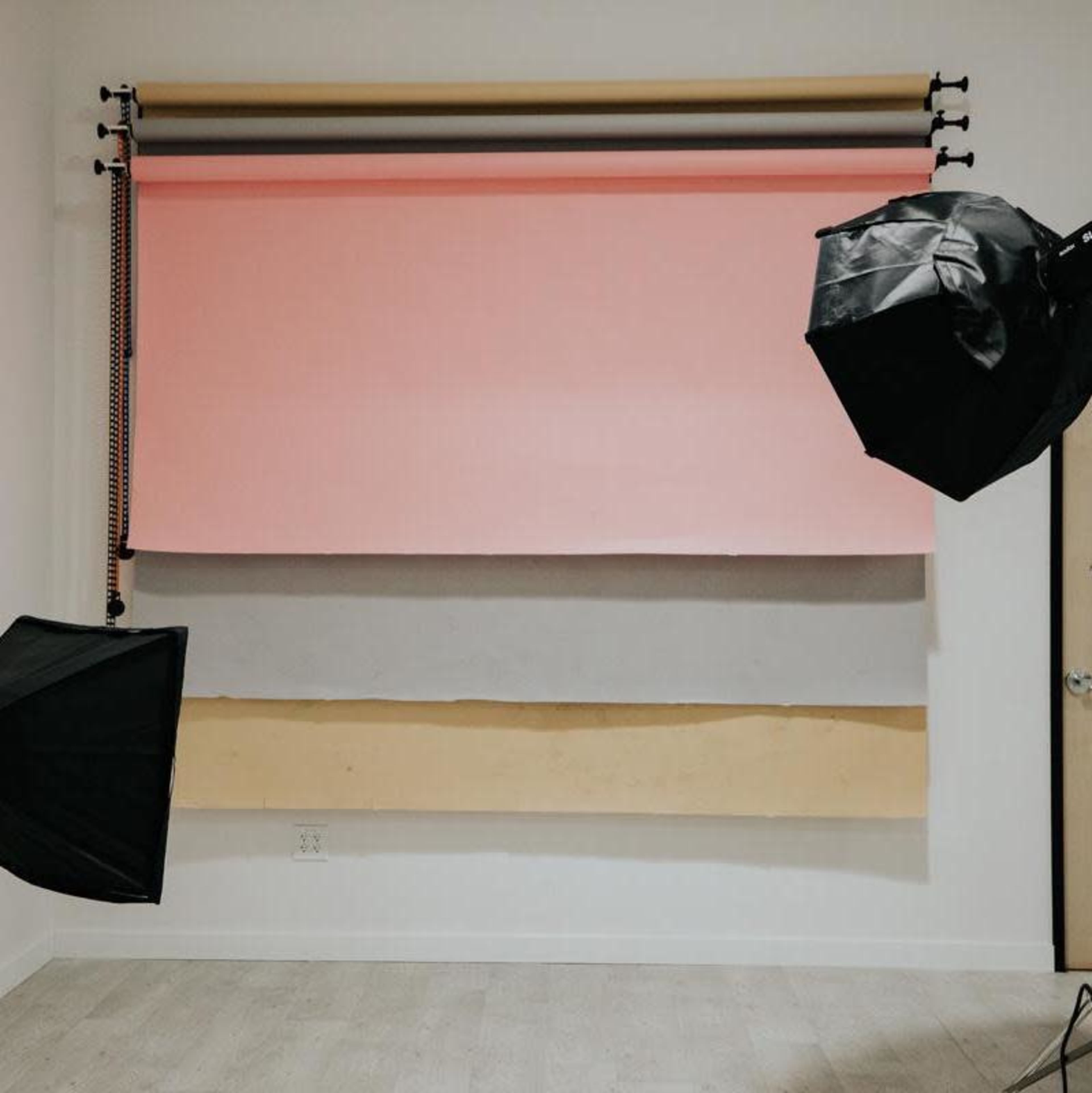 The image shows a photography studio setup with three backdrop rolls in pink, gray, and beige, flanked by two softbox lights.
