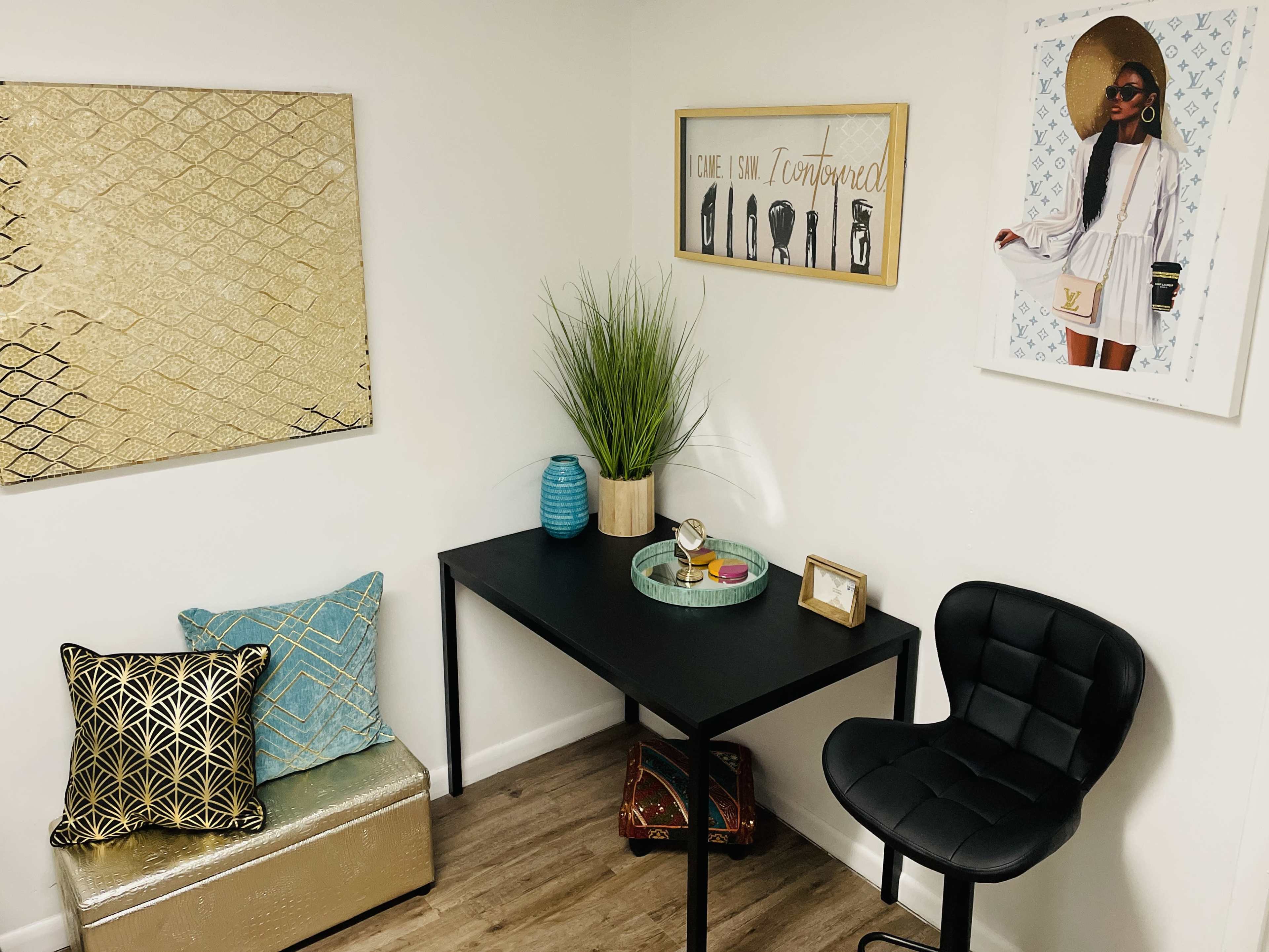The image shows a small, stylish workspace featuring a black desk, a chair, decorative pillows, and framed artwork on the walls.