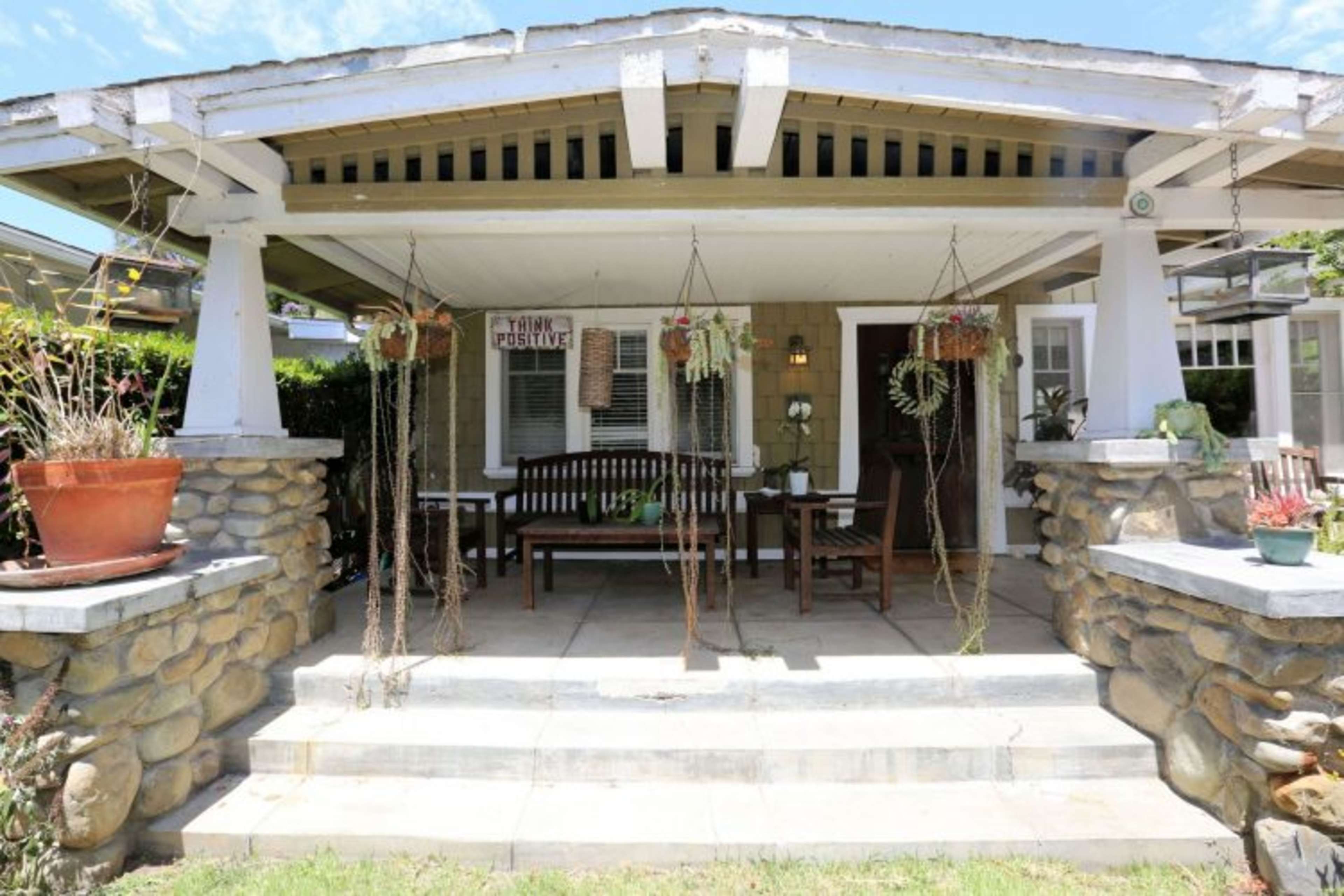 The image shows a covered porch with a wooden bench, stone columns, and hanging plants, set in a residential area.