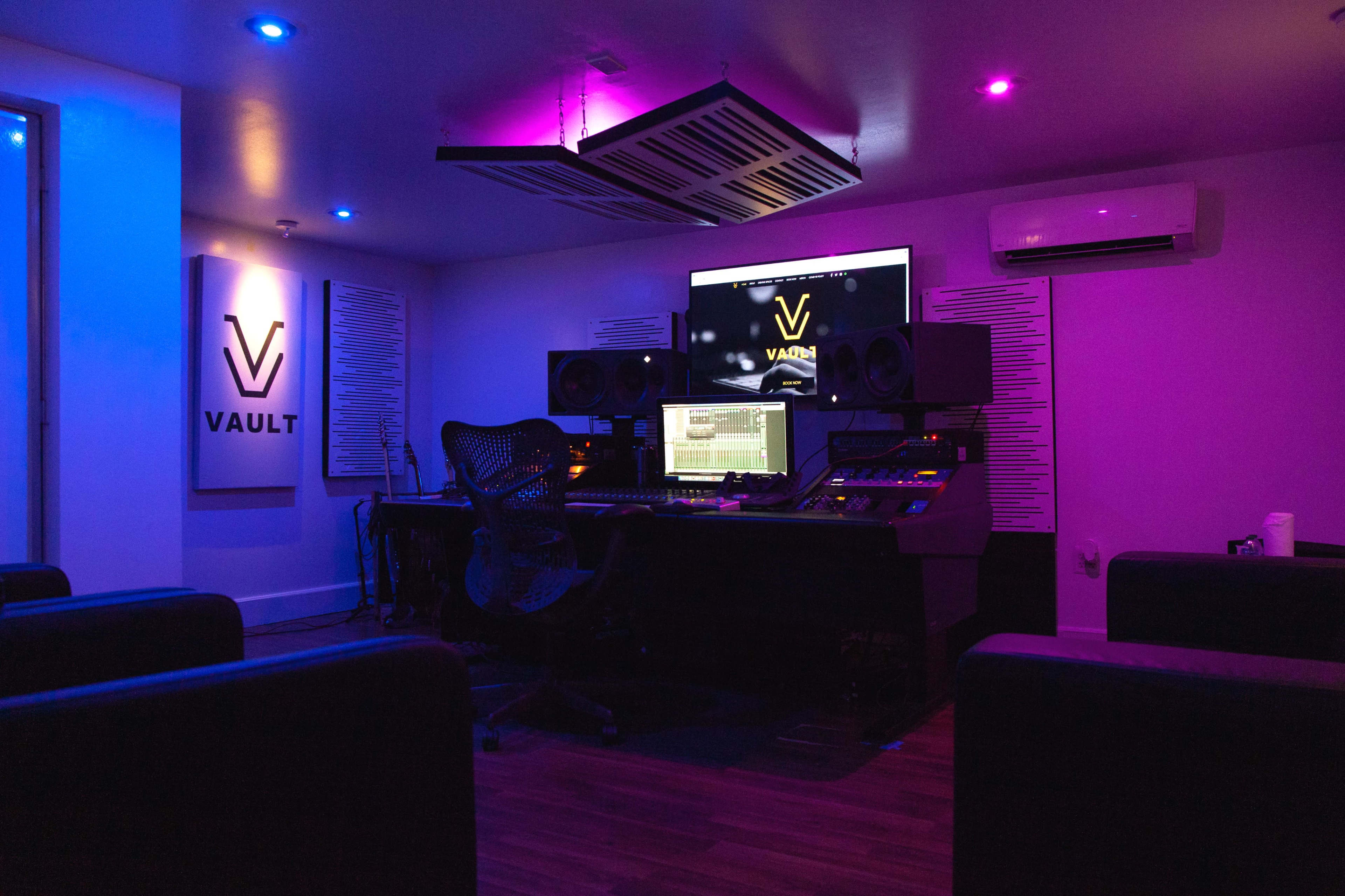A modern recording studio with a dark ambiance, featuring a large mixing console, multiple speakers, and colorful LED lighting.