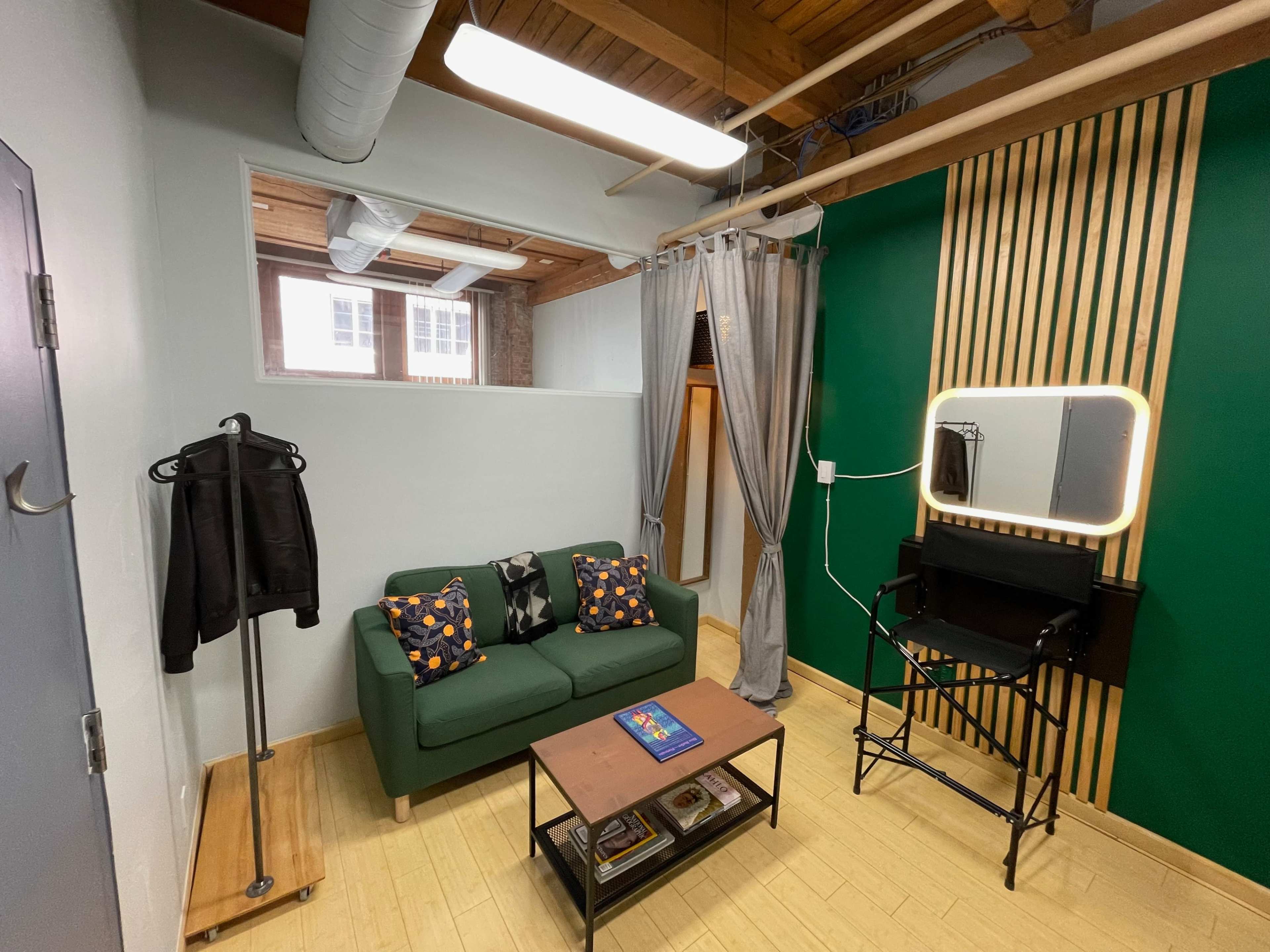 A small room with a green accent wall, a green sofa, a black director's chair, a mirror with lights, and a wooden coffee table.