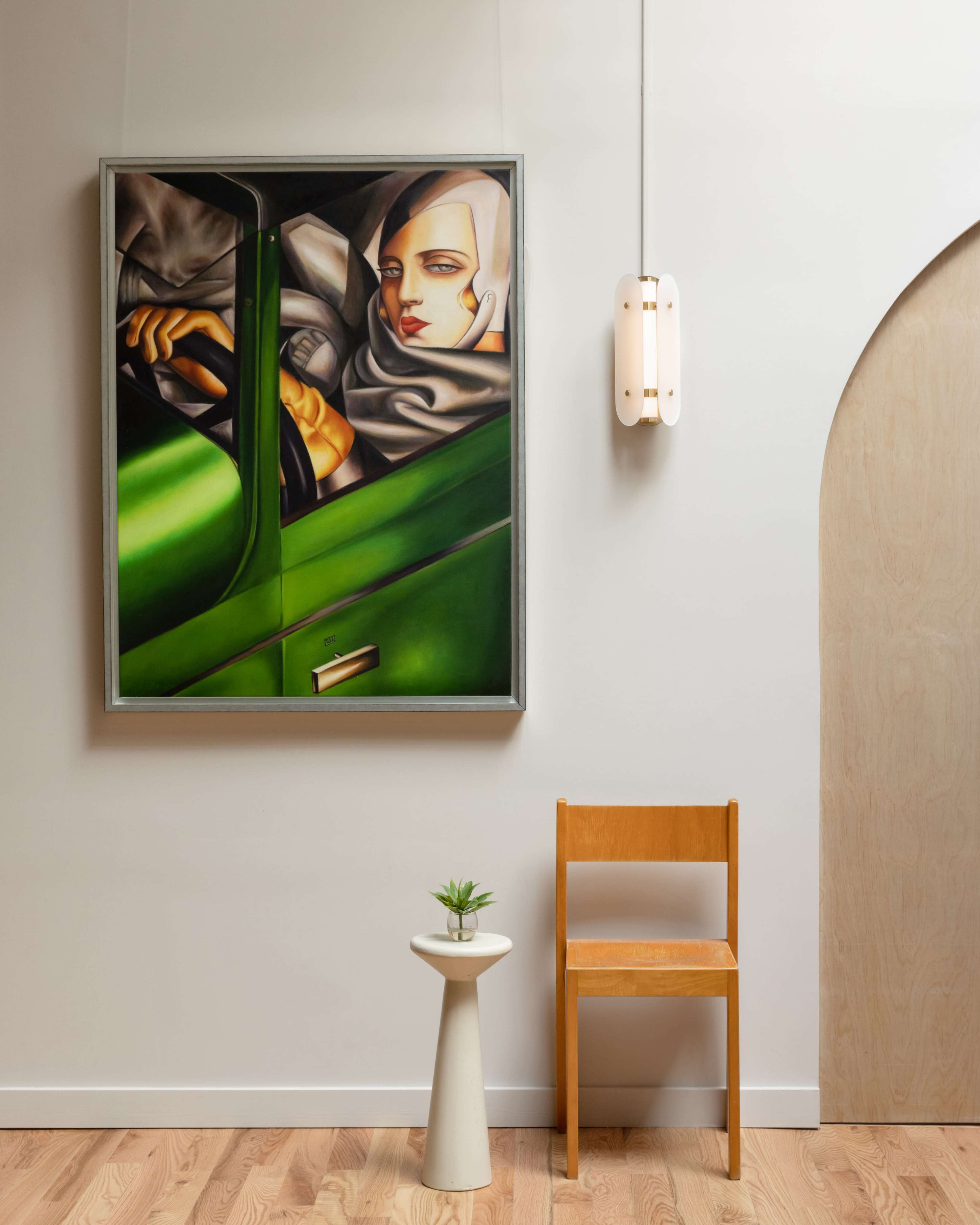 A modern interior featuring a chair, a small plant on a pedestal, and a large painting of a figure in a green car on the wall.