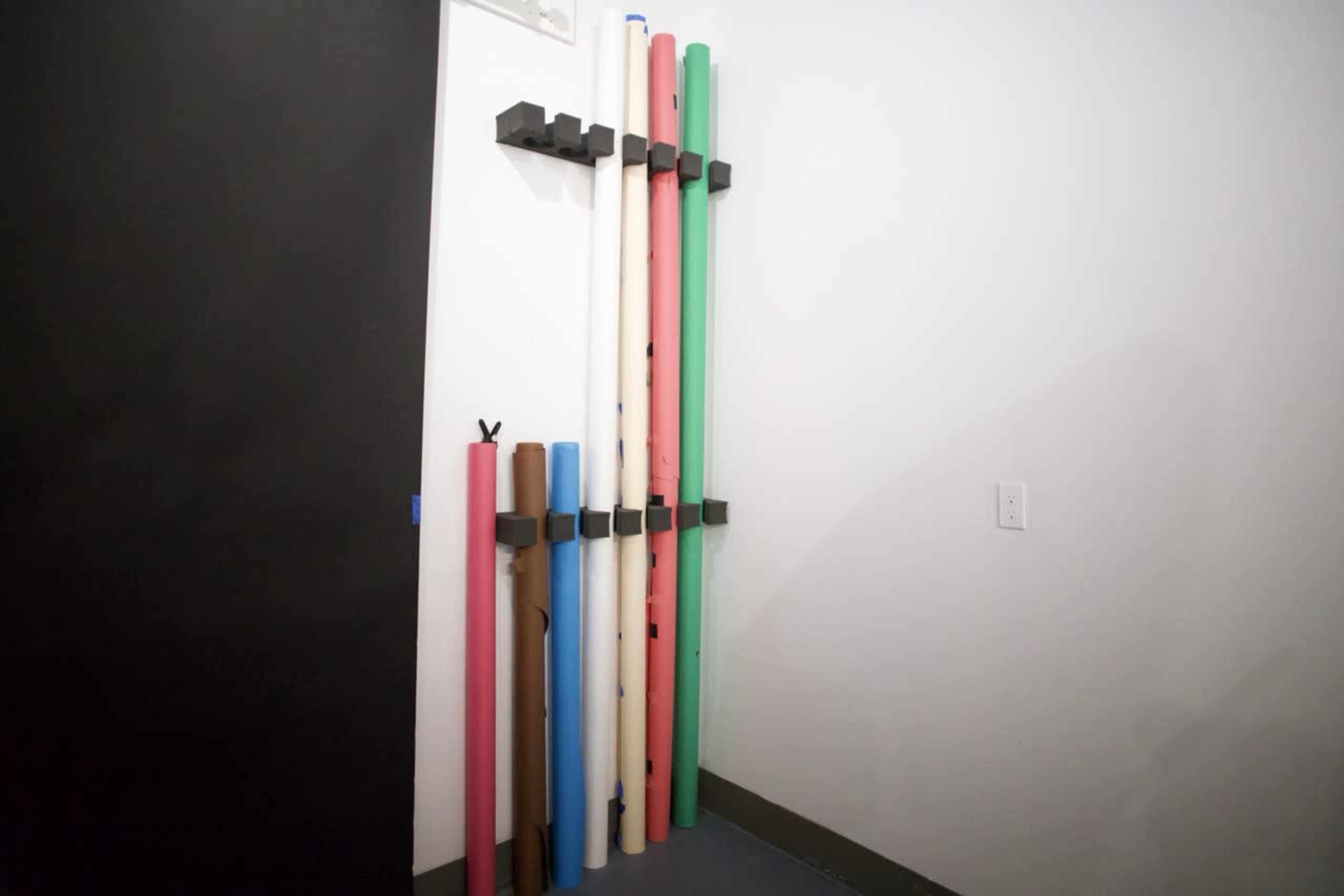 The image shows a corner of a room with several rolled-up paper or plastic sheets arranged vertically on a wall-mounted rack.