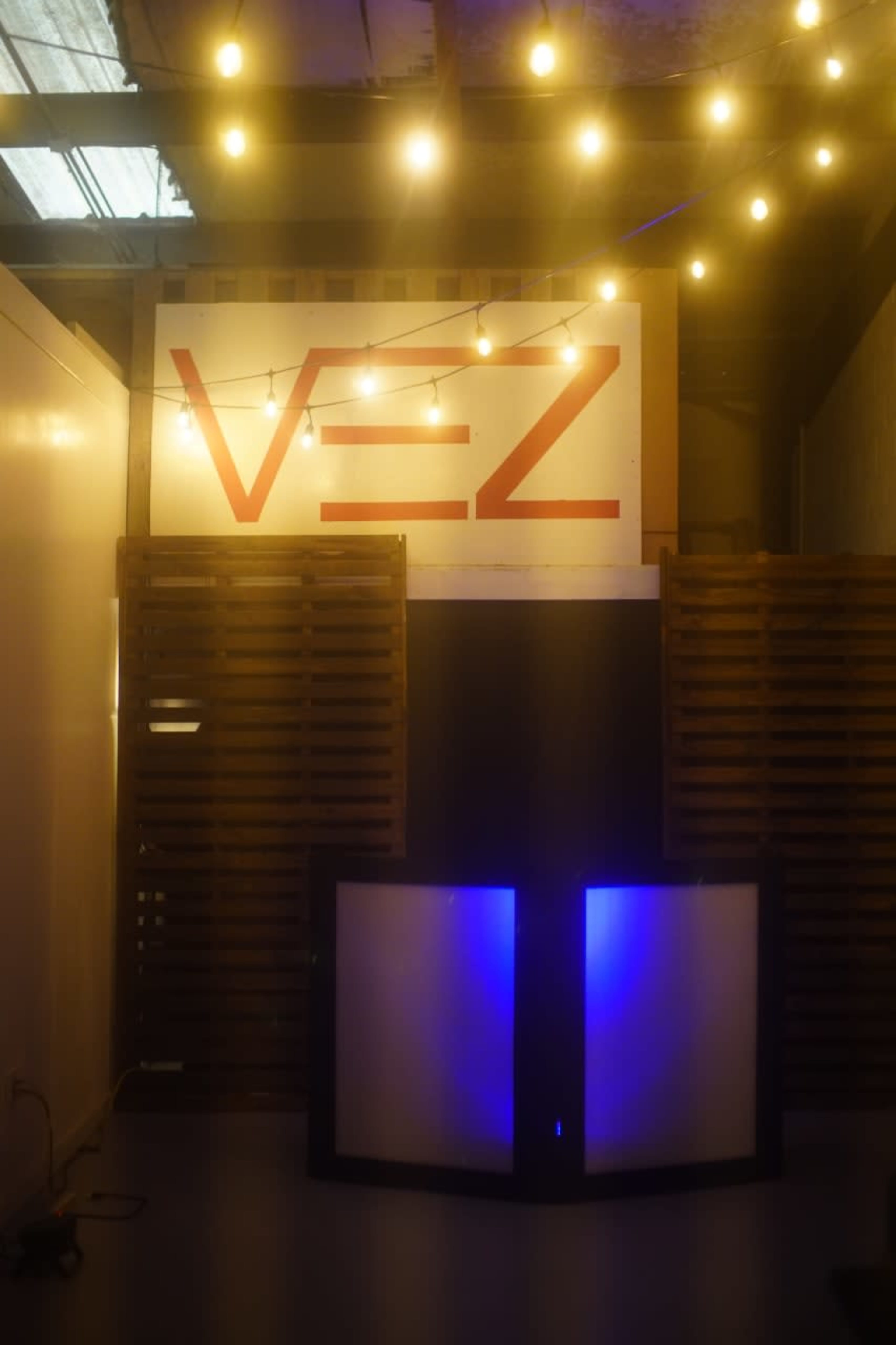 A dimly lit event space featuring a large sign with the text "V-E-Z" and a backdrop of wooden pallets, accompanied by string lights overhead.