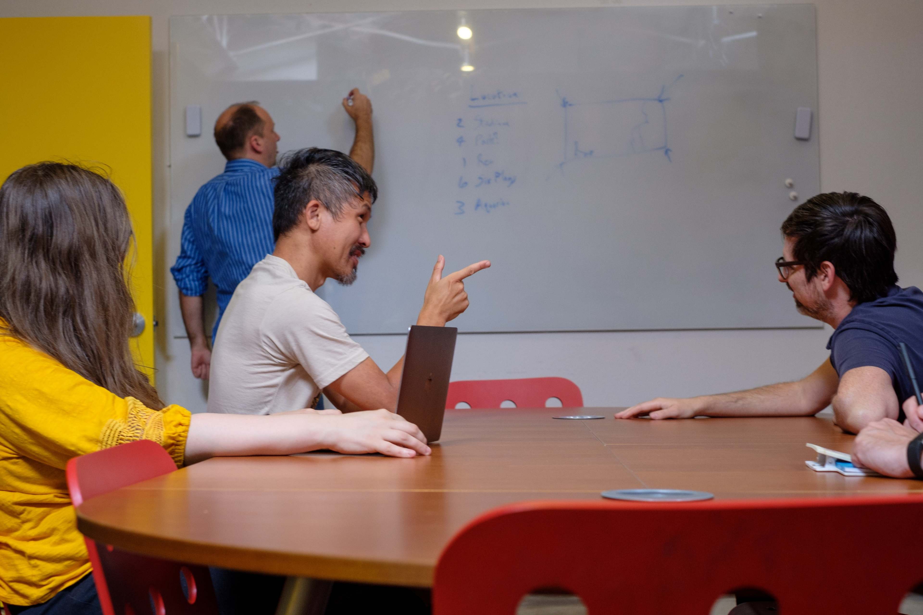 A group of people sits around a conference table, with one person writing on a whiteboard while others engage in discussion.