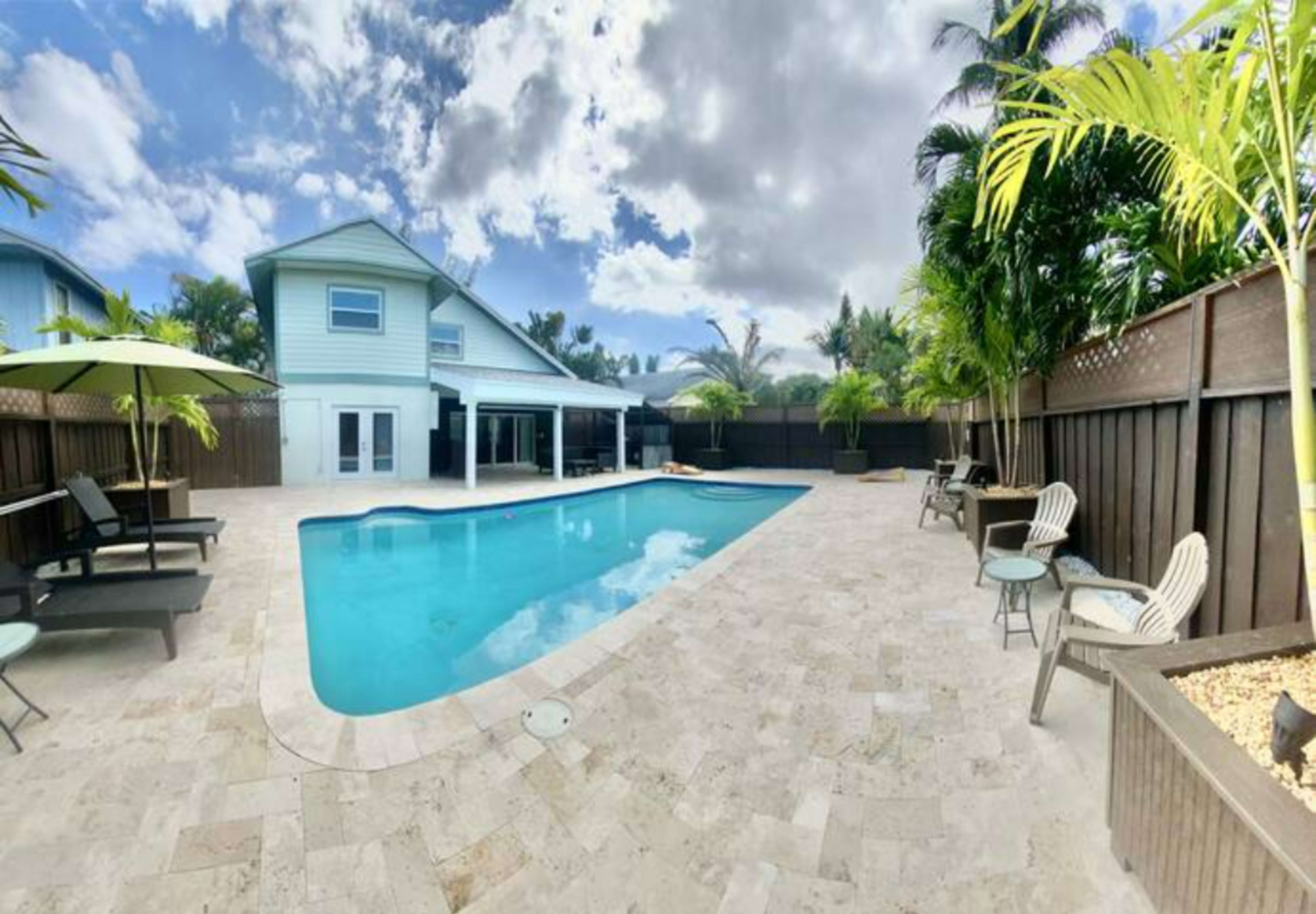 A rectangular swimming pool surrounded by lounge chairs and tropical plants, with a two-story house in the background under a partly cloudy sky.