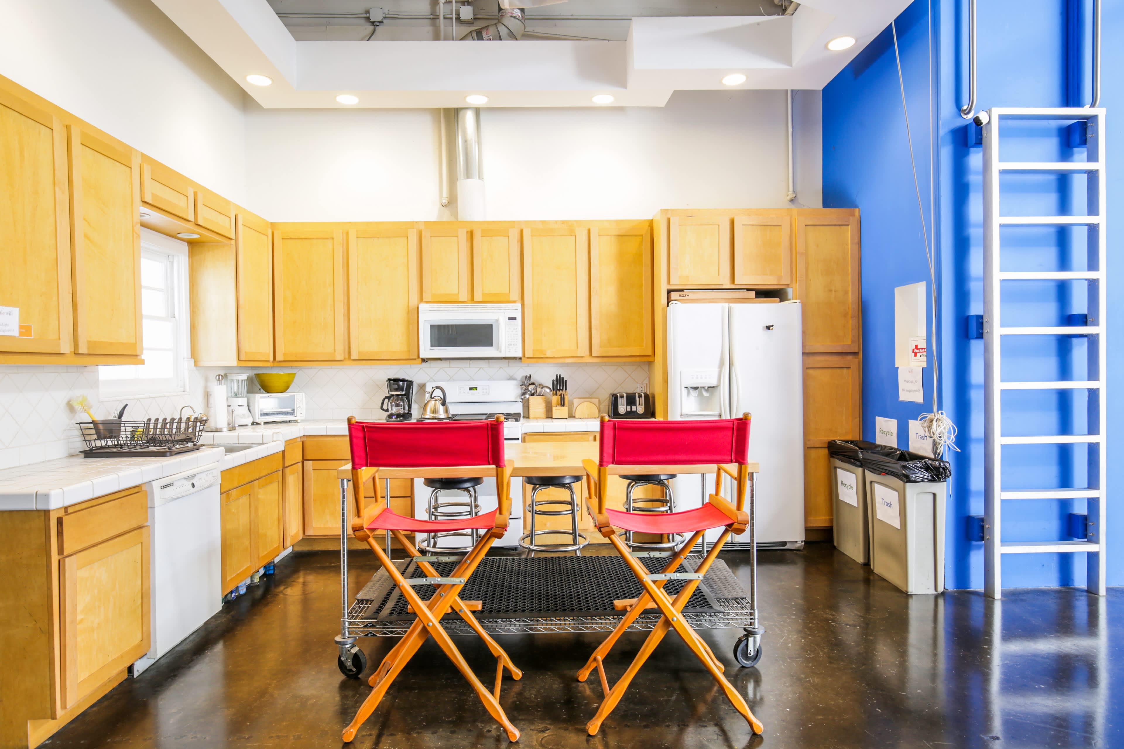 A modern kitchen features light wood cabinetry, stainless steel appliances, and two red director's chairs positioned in front of a quartz countertop.
