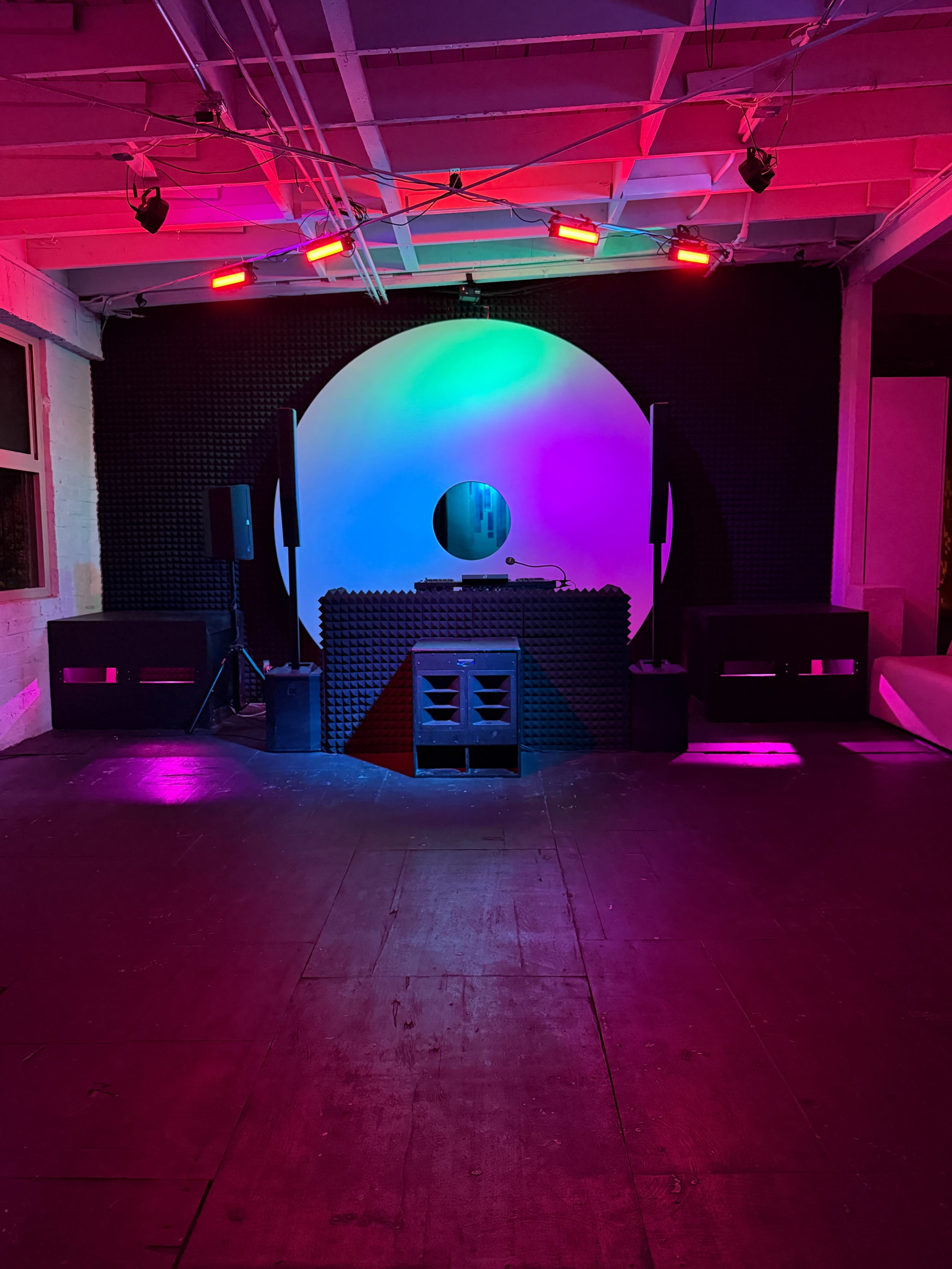 The image shows a music performance space featuring a DJ booth with colorful lighting and a large circular backdrop.