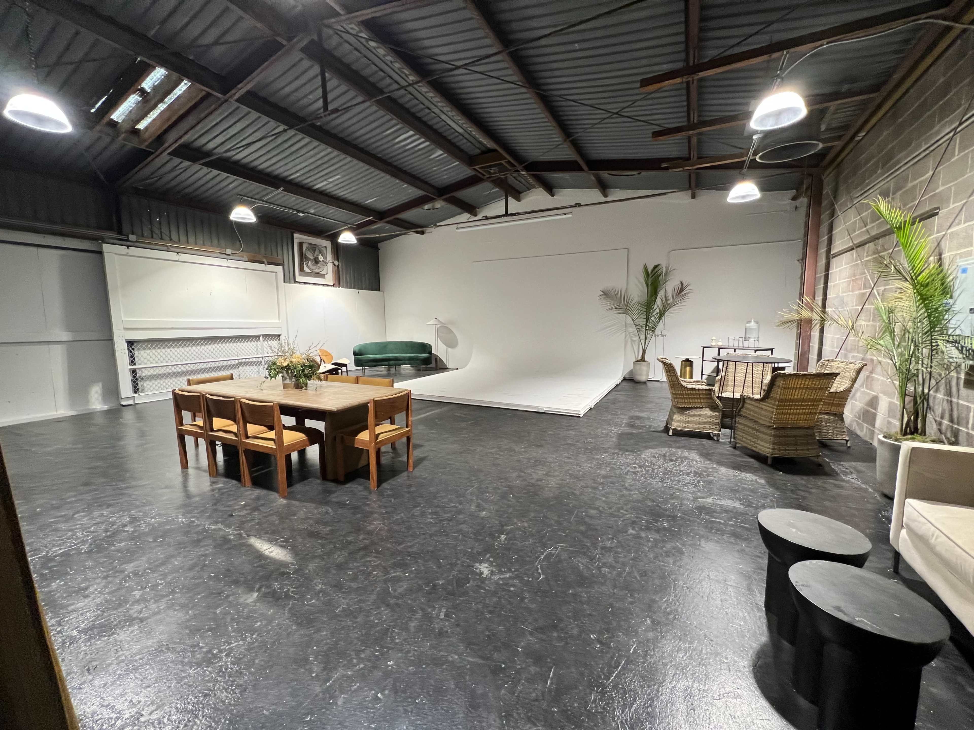 A spacious industrial-style room with a large empty backdrop, a dining table, a green couch, and various seating arrangements.