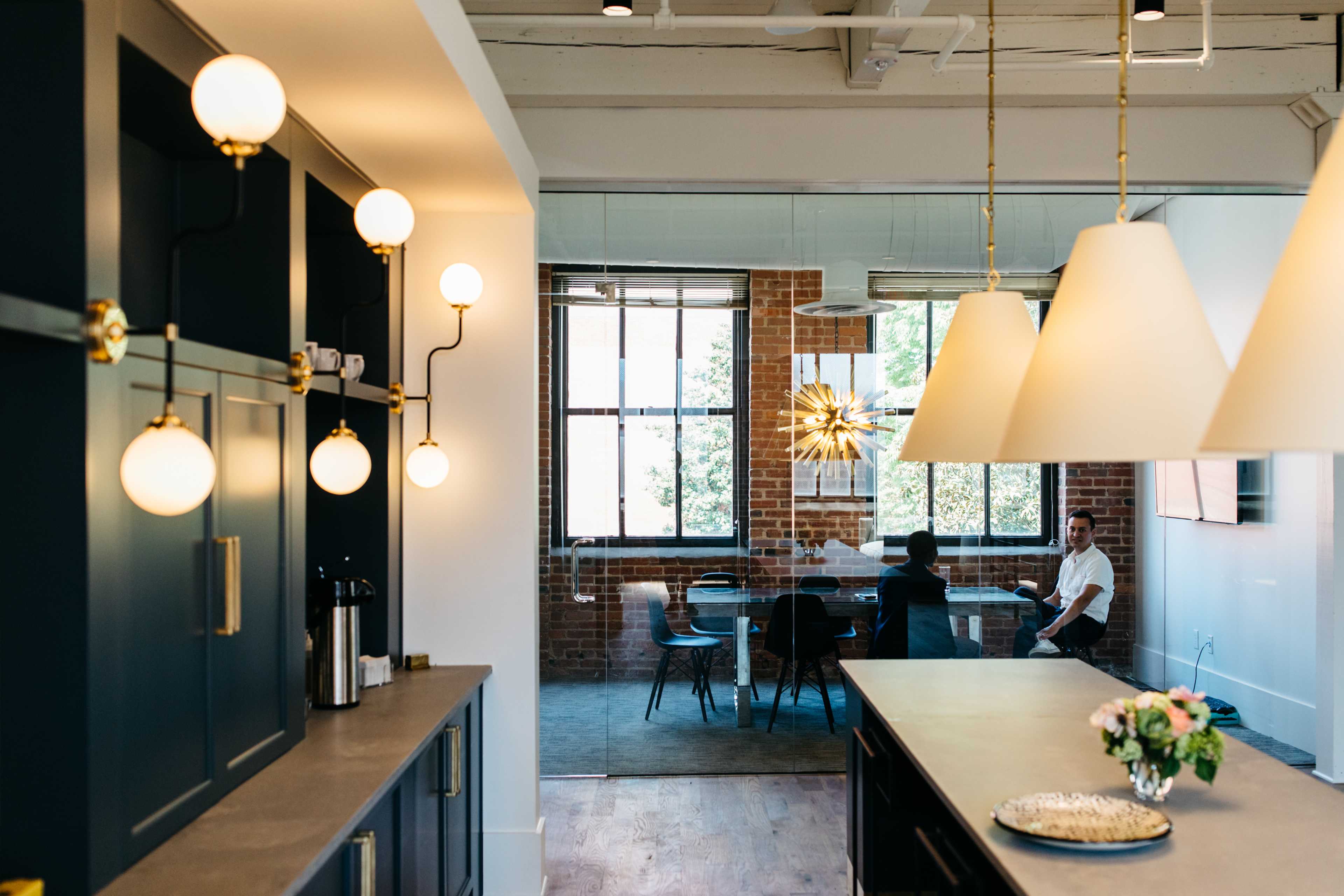 A modern office space with a kitchen area featuring round light fixtures and a dining table visible through a glass partition.