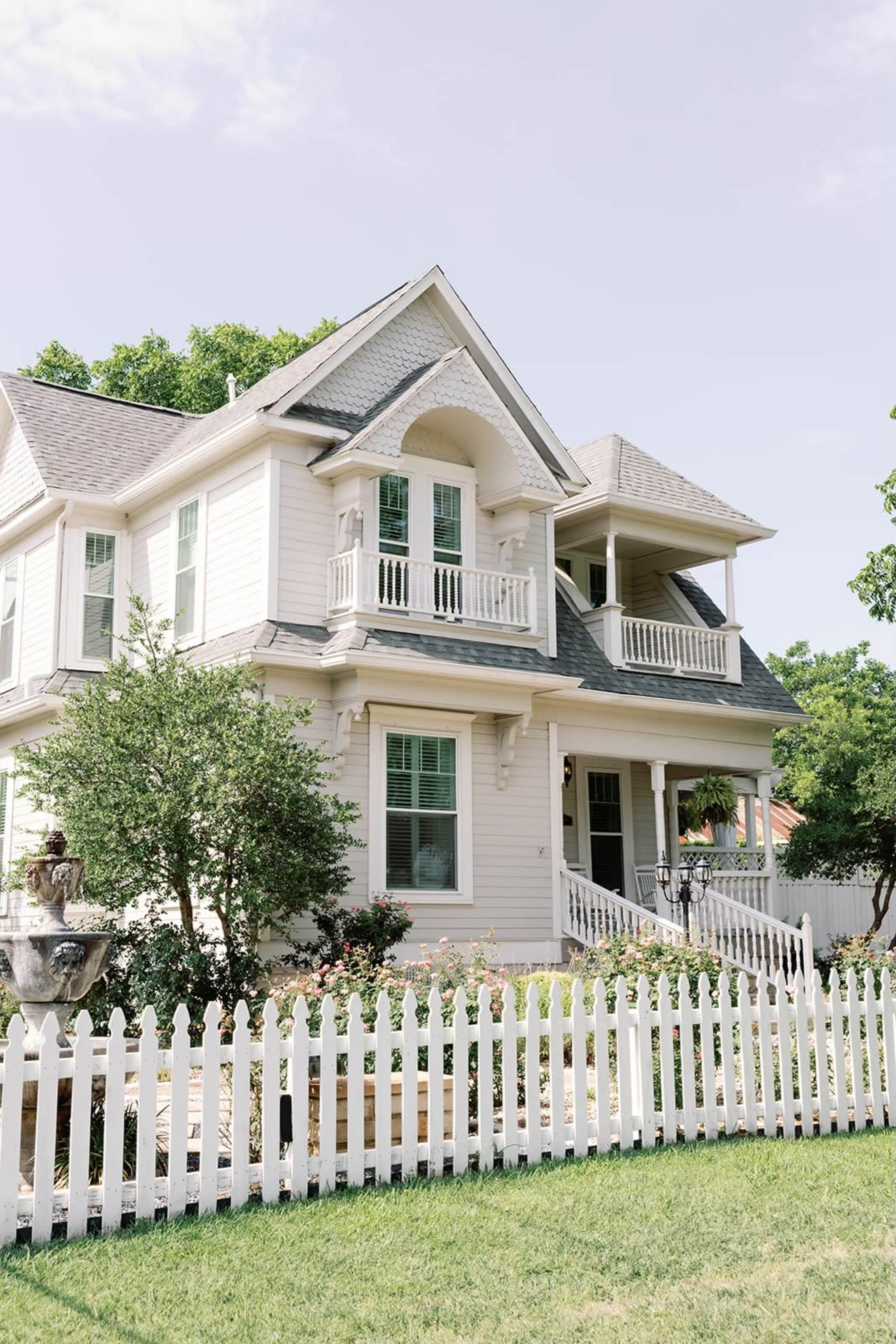 The image shows a two-story house with a light-colored exterior, a front porch, and a white picket fence surrounding the yard.