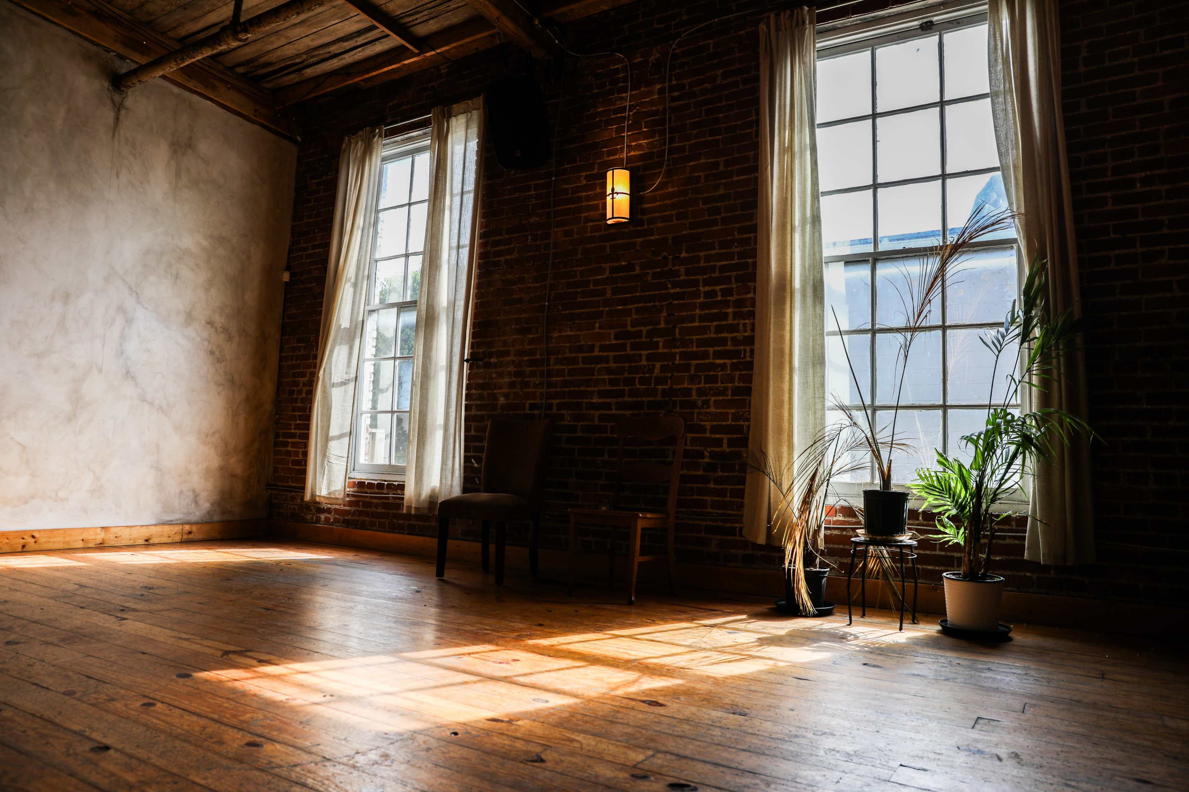 The image shows a sunlit room with brick walls, wooden floors, two large windows, a chair, and indoor plants.